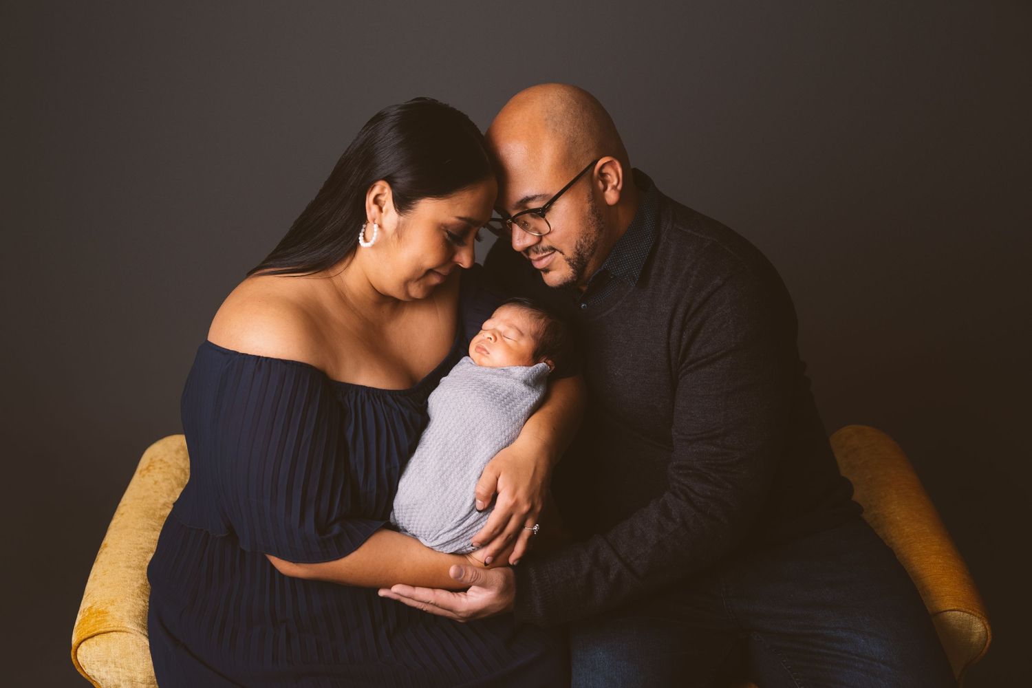 Intimate family moment with parents cradling their sleeping newborn against dark backdrop.
