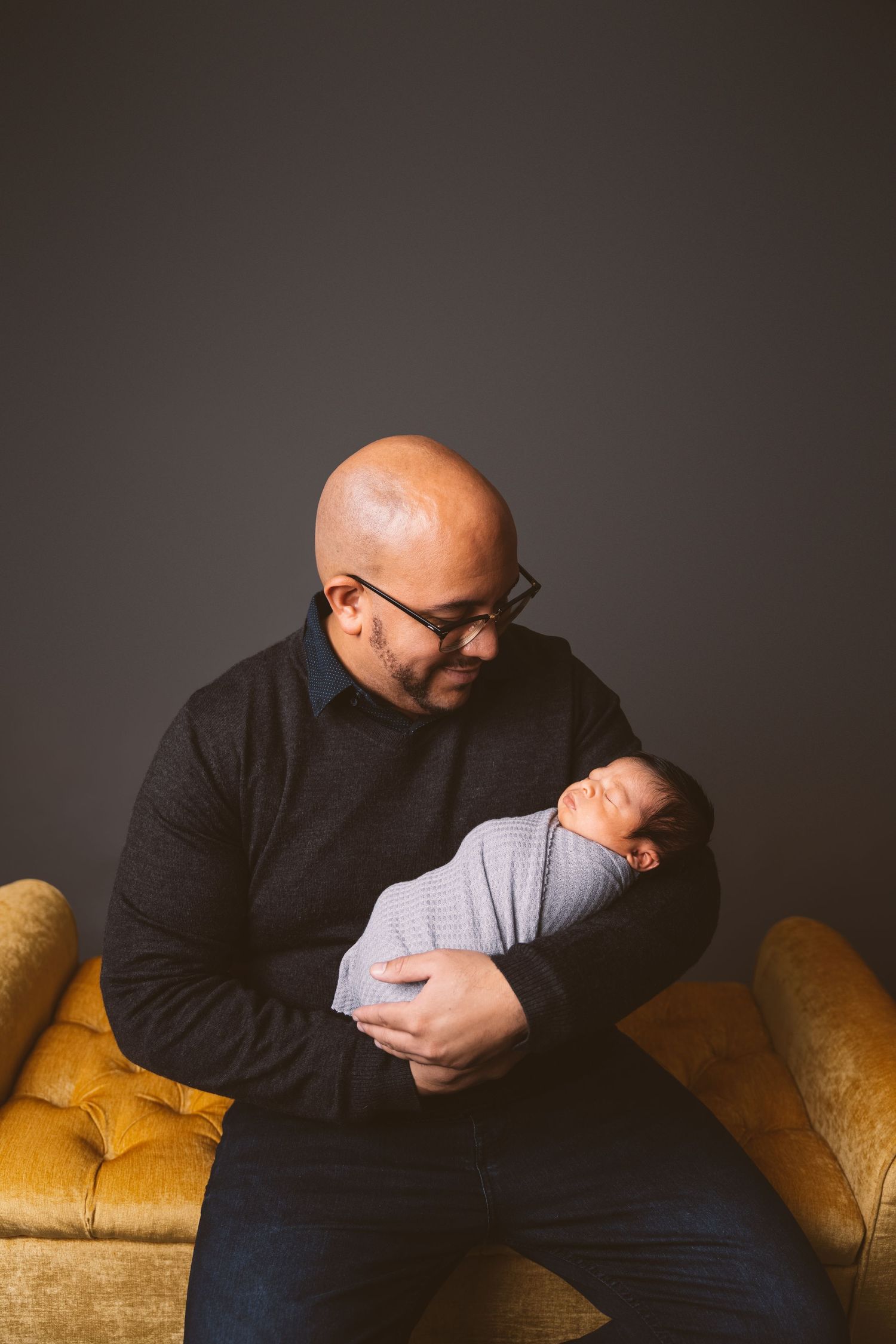 Parent tenderly holding sleeping newborn on yellow couch against dark background.