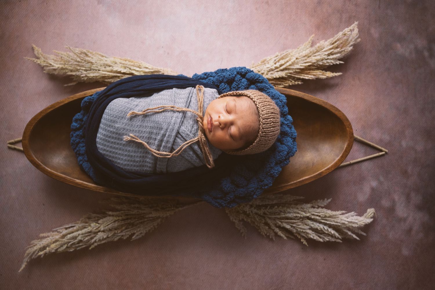 Artistic newborn portrait with baby sleeping in wooden bowl surrounded by dried wheat stalks.