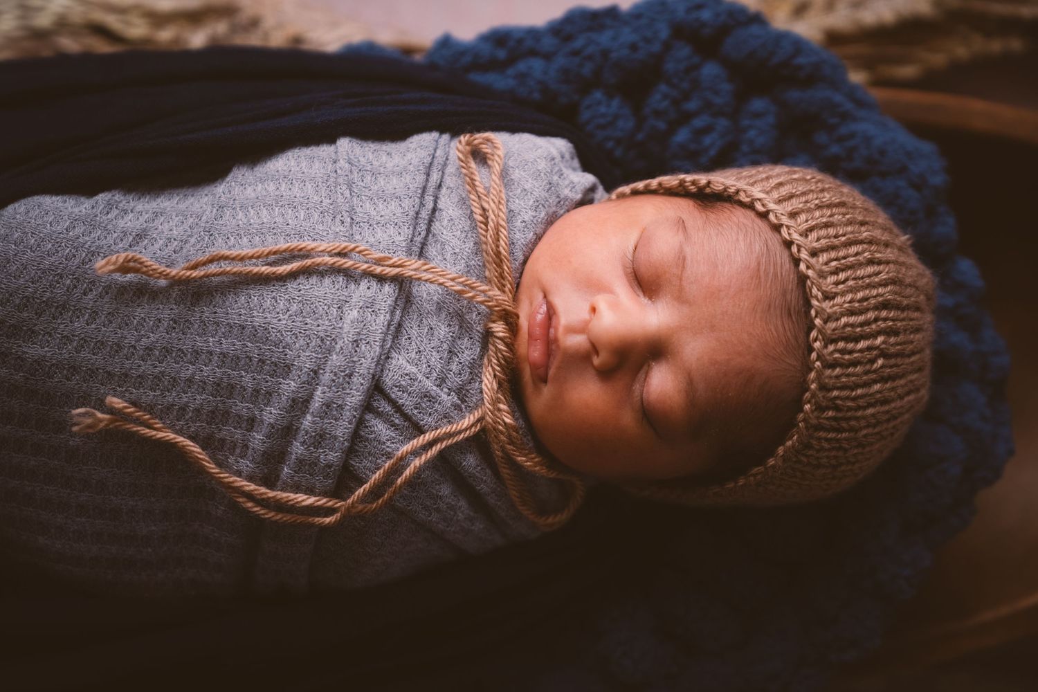 Sleeping newborn wearing knit beanie and denim in cozy blue blanket during professional photoshoot.