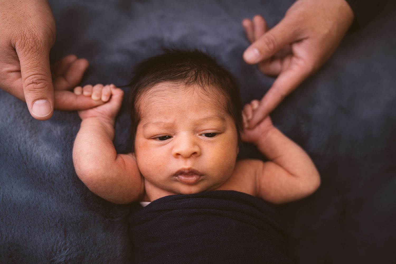 Alert newborn baby resting on dark blue blanket with supporting hands.