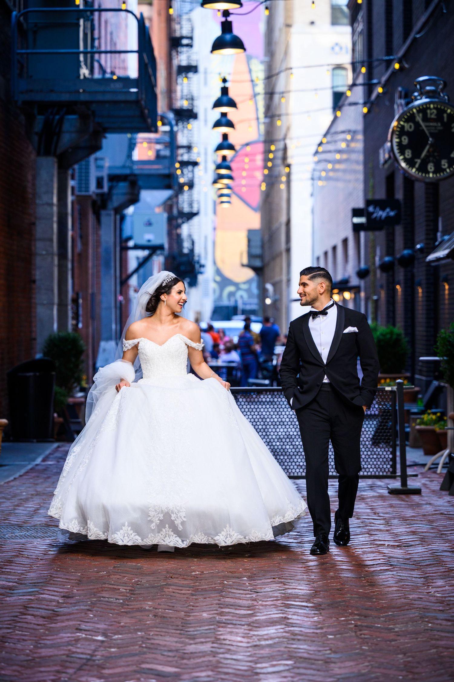 A wedding couple walks along a romantic cobblestone street with hanging lights and historic buildings in Boston.