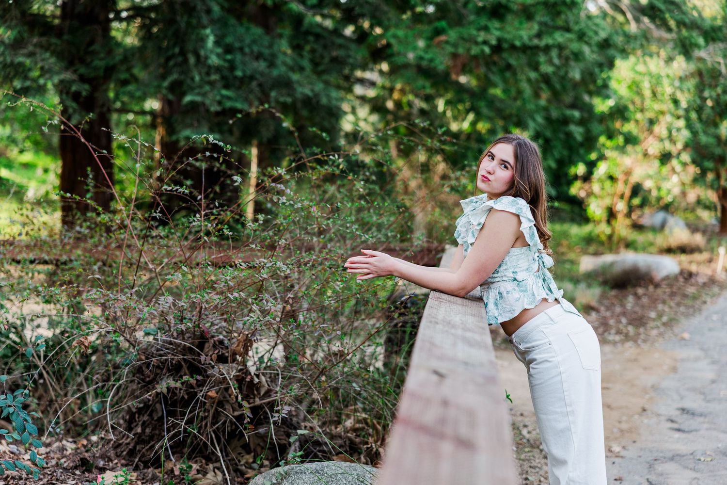 Someone wearing a denim vest and cream pants leans against a stone wall on a forest path in summer.