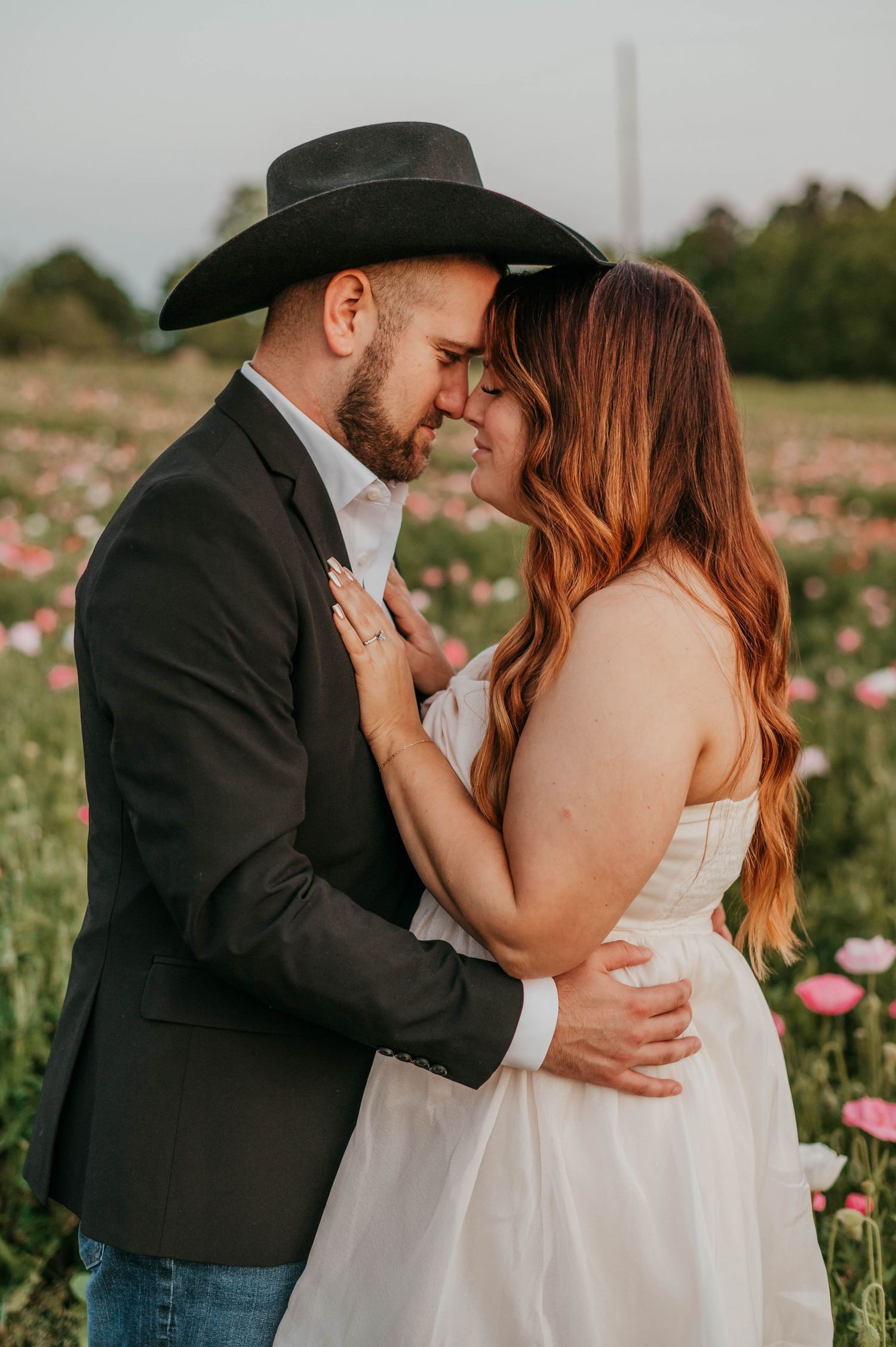 A romantic couple embraces in a field of pink wildflowers during sunset, wearing wedding attire and a cowboy hat.