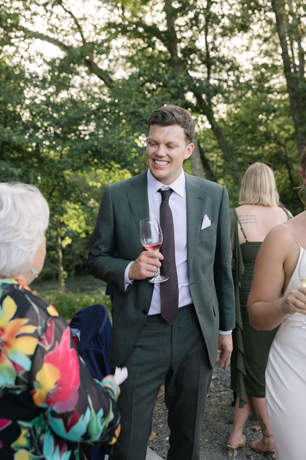 Wedding guest wearing dark green suit holds wine glass at outdoor reception with trees in background.