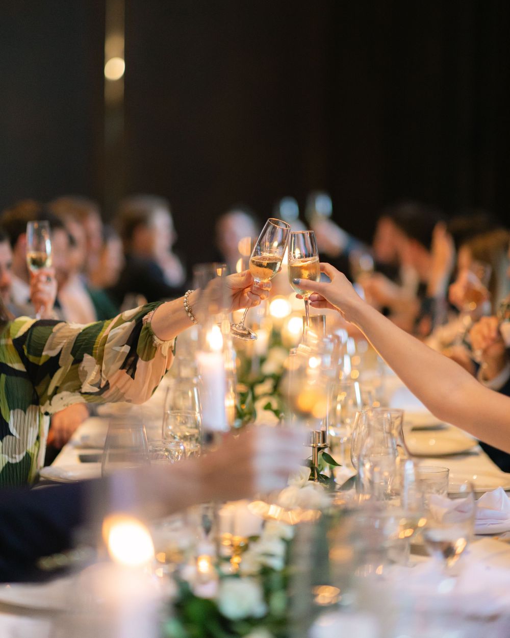 Guests raise wine glasses for a candlelit wedding toast at an elegantly decorated dinner table with greenery centerpieces.