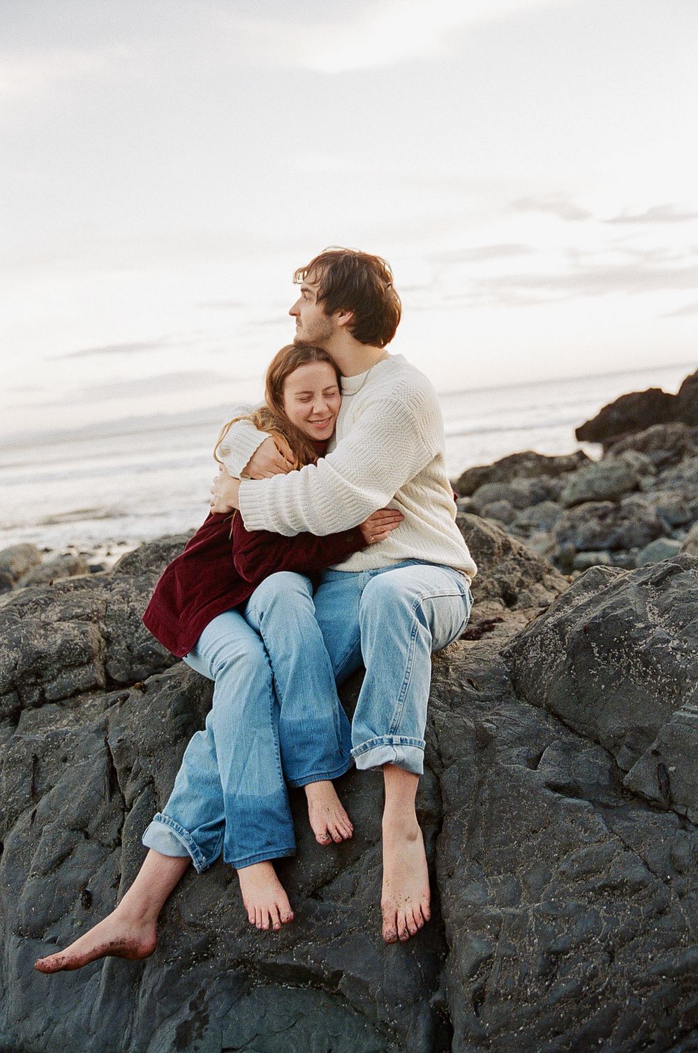A couple embraces while sitting barefoot on rocky coastal outcrop at sunset, wearing casual denim and cream sweater.