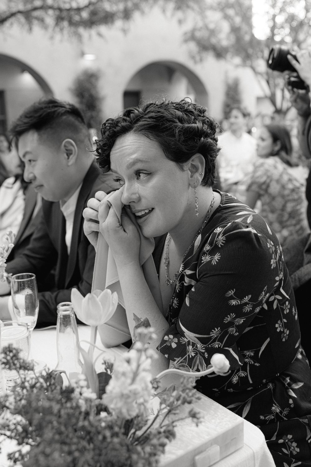 A candid black and white photo of a person in a floral dress laughing while dining outdoors at an elegant event.