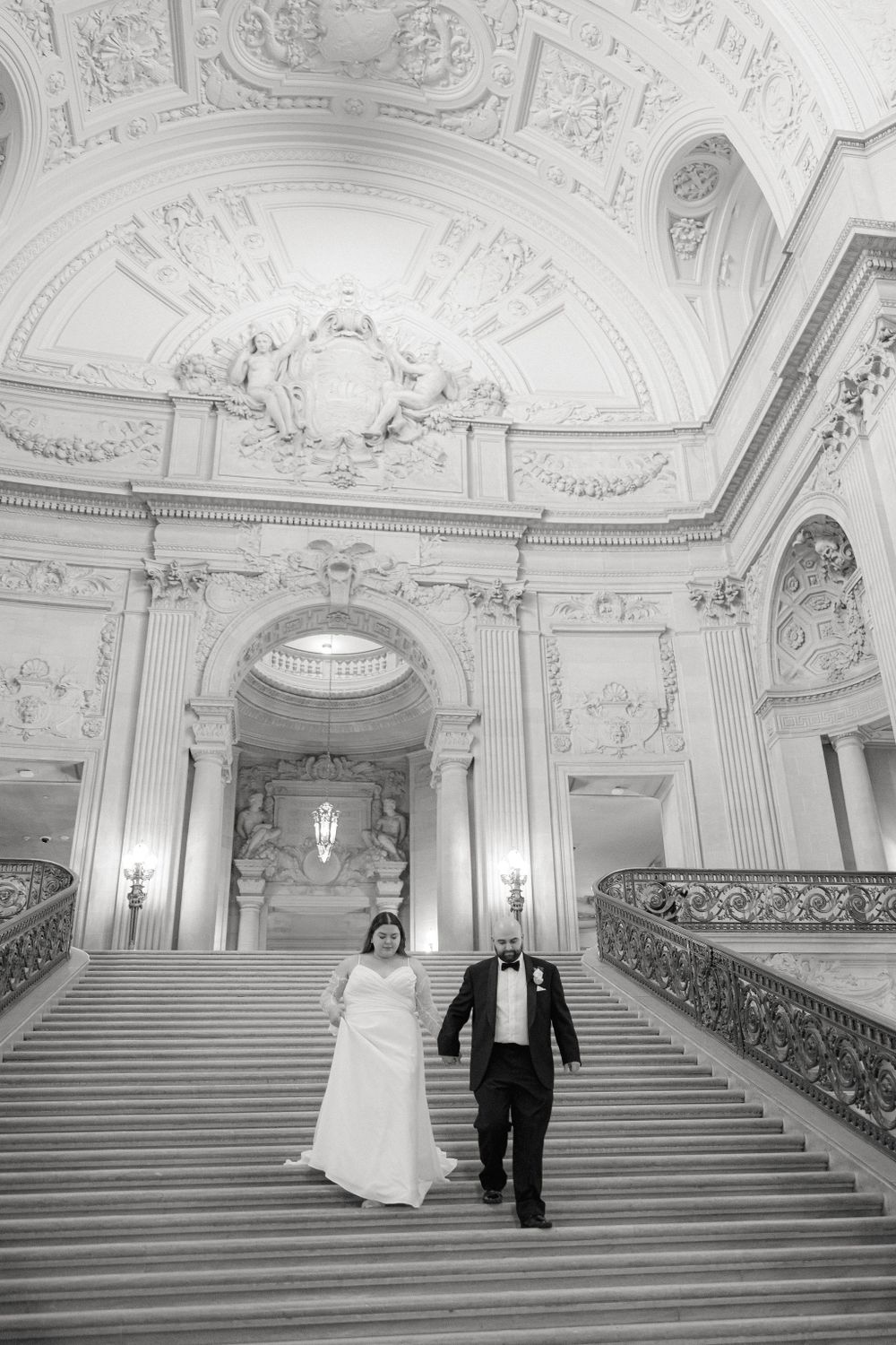Couple walks down grand marble staircase in ornate historic building with decorative white baroque interior architecture.