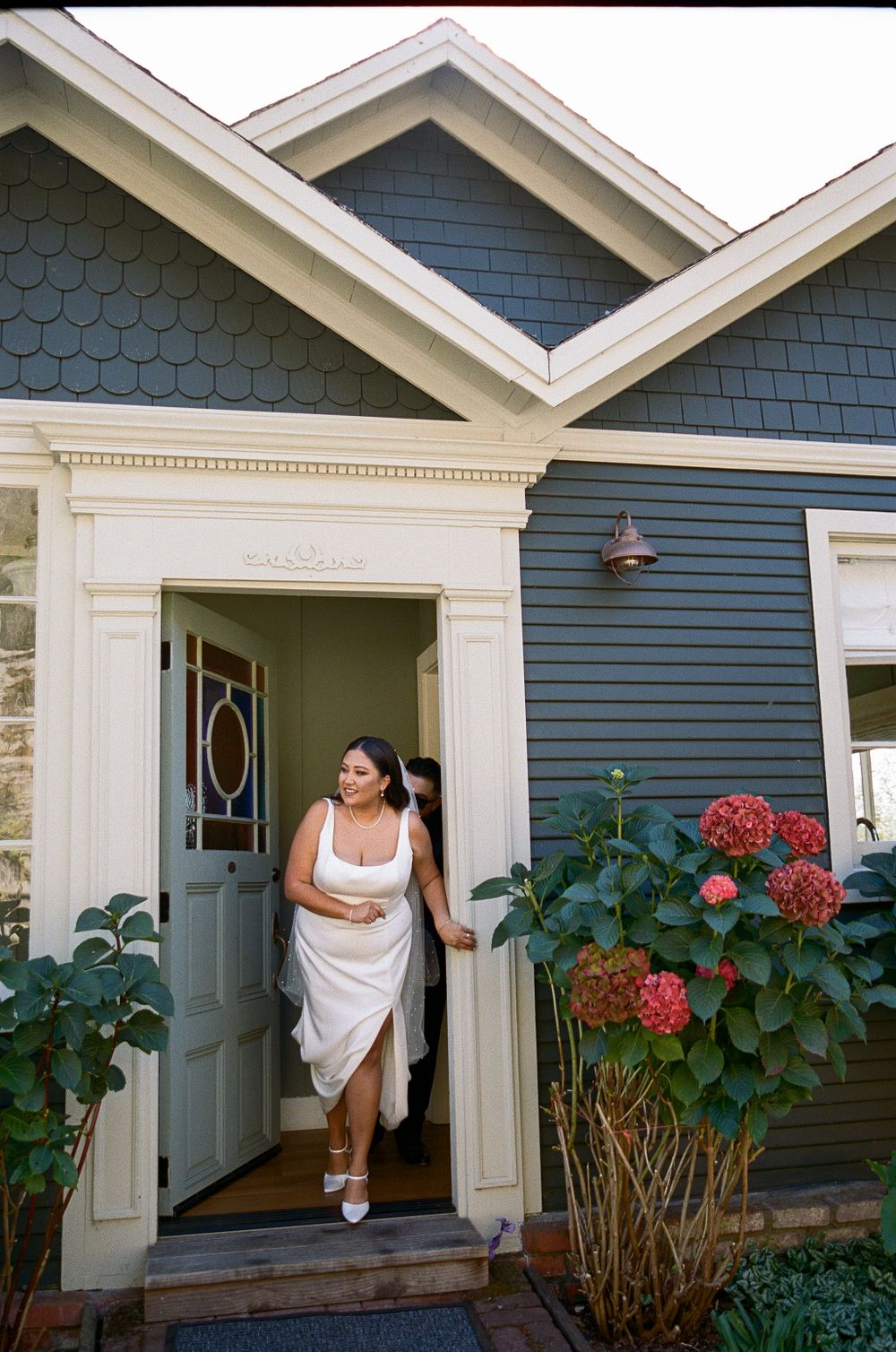 A grey-blue craftsman style house with white trim features a mint green door and blooming coral hydrangeas by the entrance.