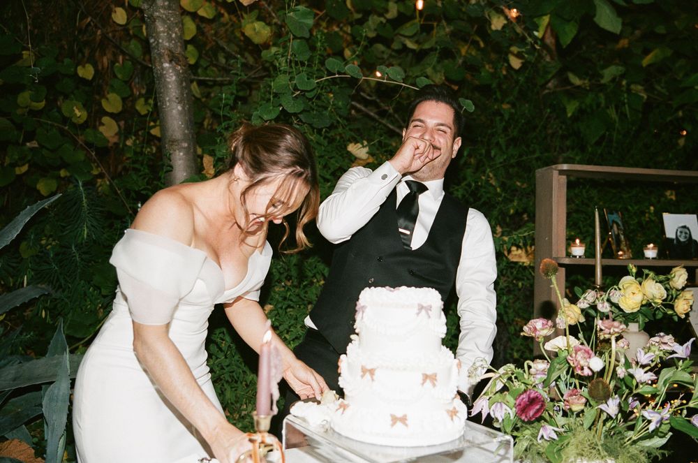 A couple laughs while cutting their wedding cake together at an outdoor garden reception.