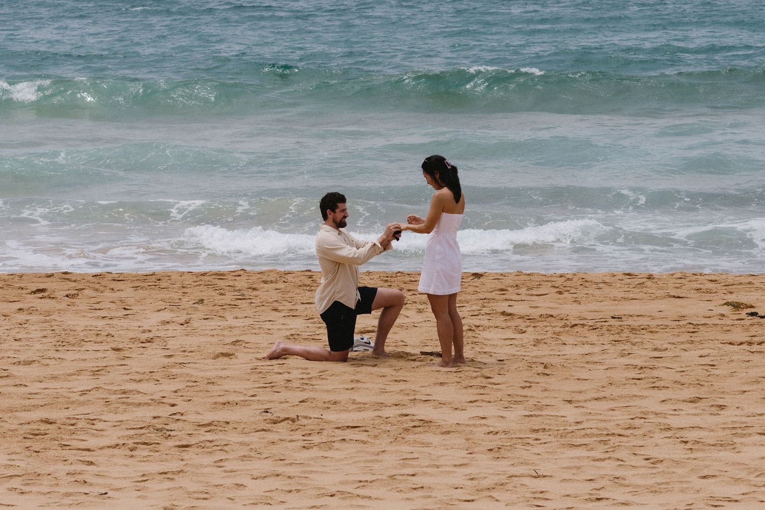 A proposal moment captured on a sandy beach with ocean waves in the background.