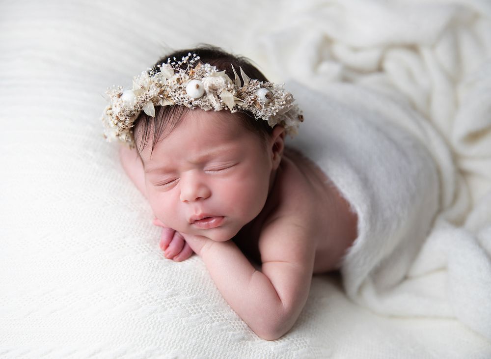 Peaceful sleeping newborn wearing delicate white floral crown rests on soft white fabric.