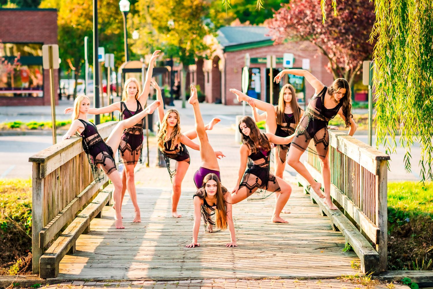 Dancers strike playful poses on a wooden bridge surrounded by autumn foliage.