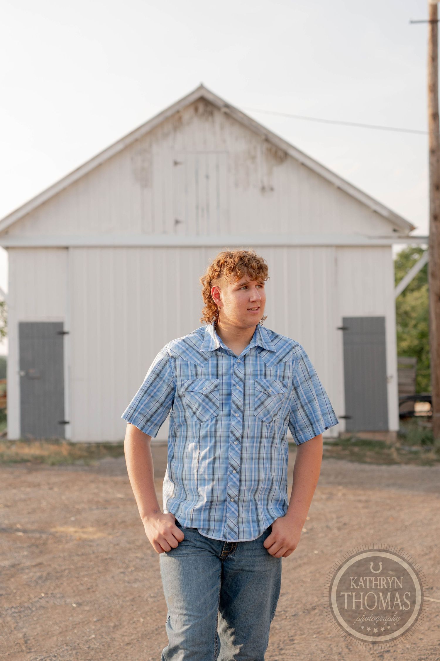 A portrait in front of a classic white barn wearing a blue plaid shirt and jeans.