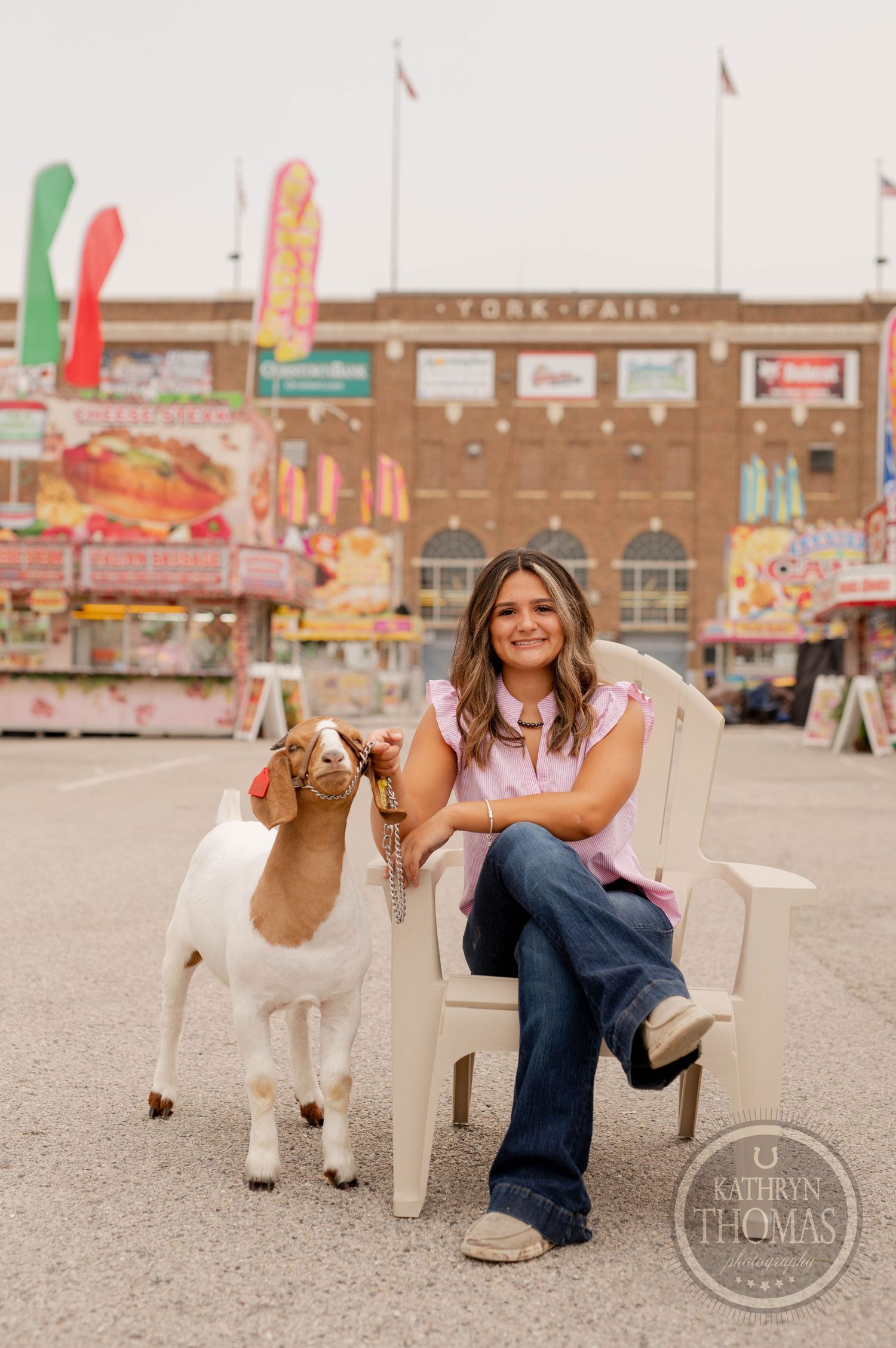 White goat poses next to someone seated in a white plastic chair at a fair with concession stands behind.