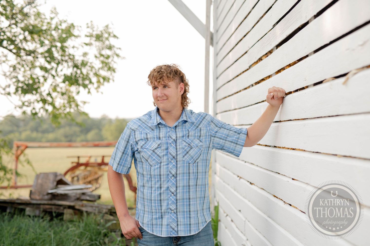 A relaxed portrait pose leaning against a rustic white barn wall in casual western attire.