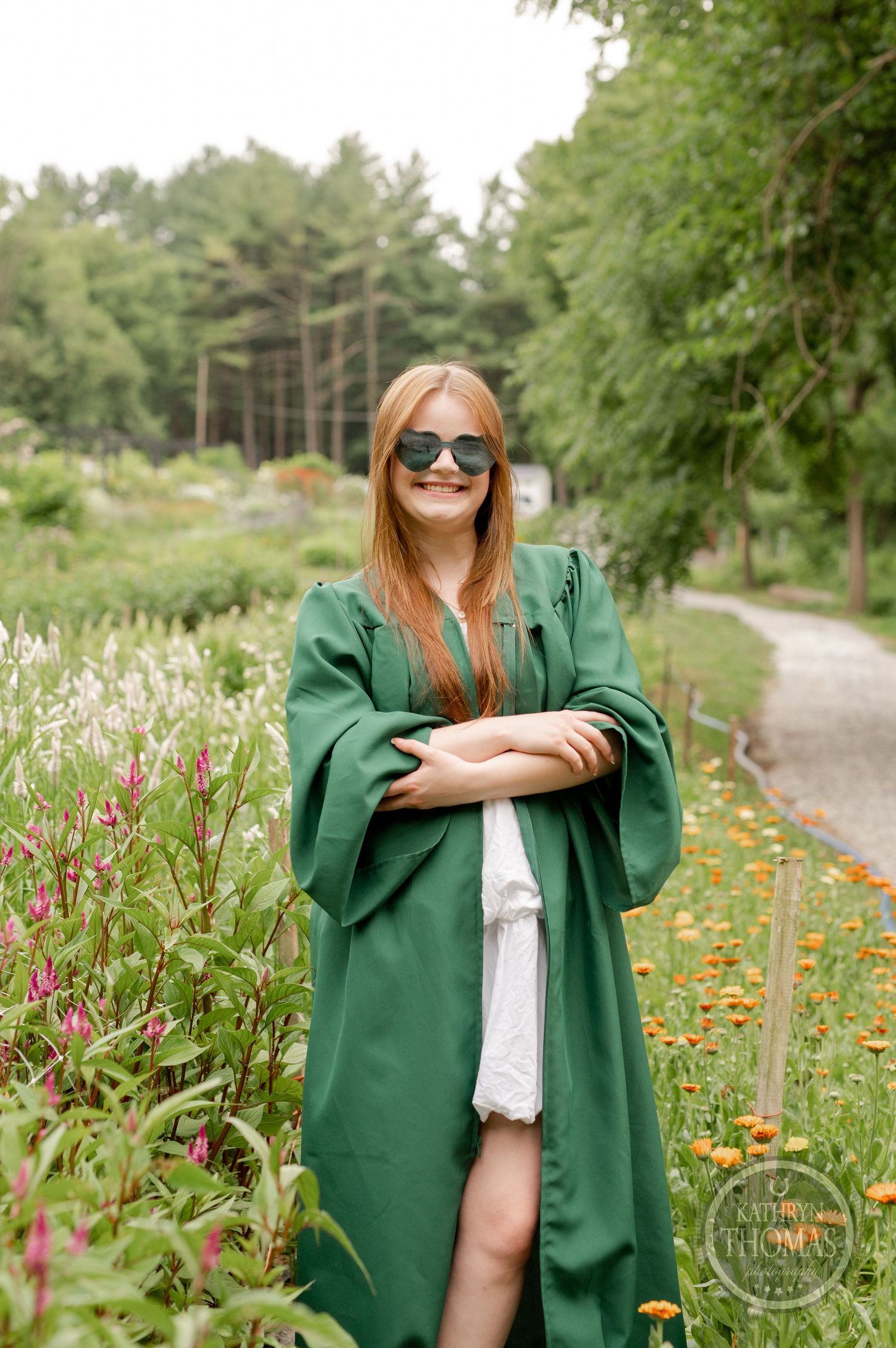 Graduate wearing sunglasses and green graduation gown poses among garden flowers.