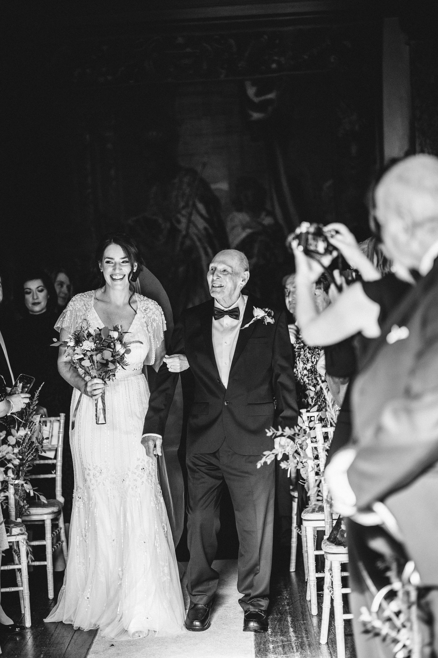 A bride and escort walk down the aisle during a wedding ceremony as guests look on and take photos.