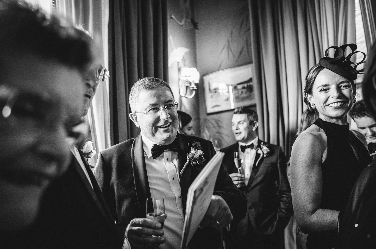 Black and white photograph of people at a social gathering with formal attire and elegant decor in the background.