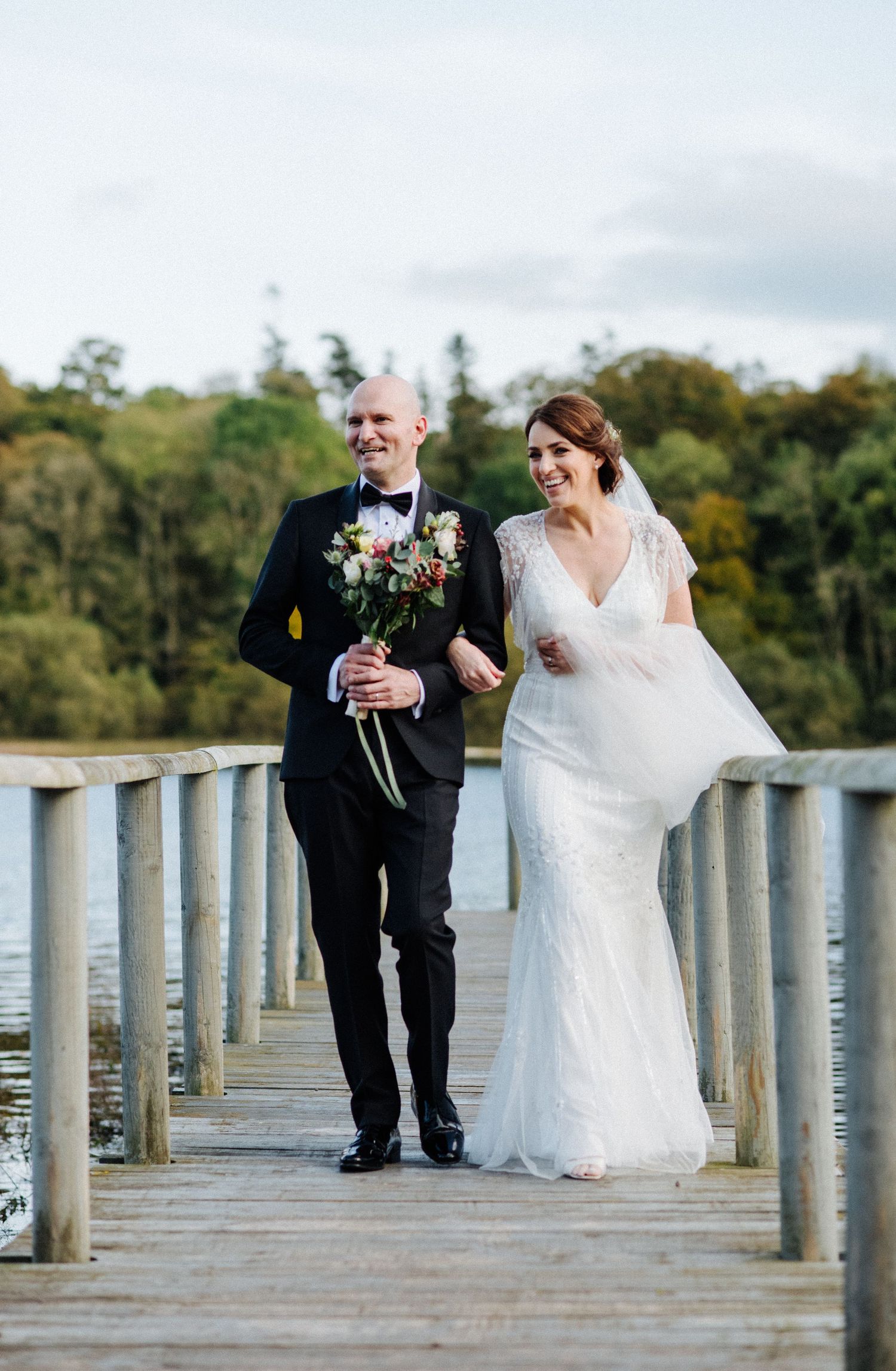 A couple in wedding attire walks arm-in-arm along a wooden dock over water surrounded by autumn trees.