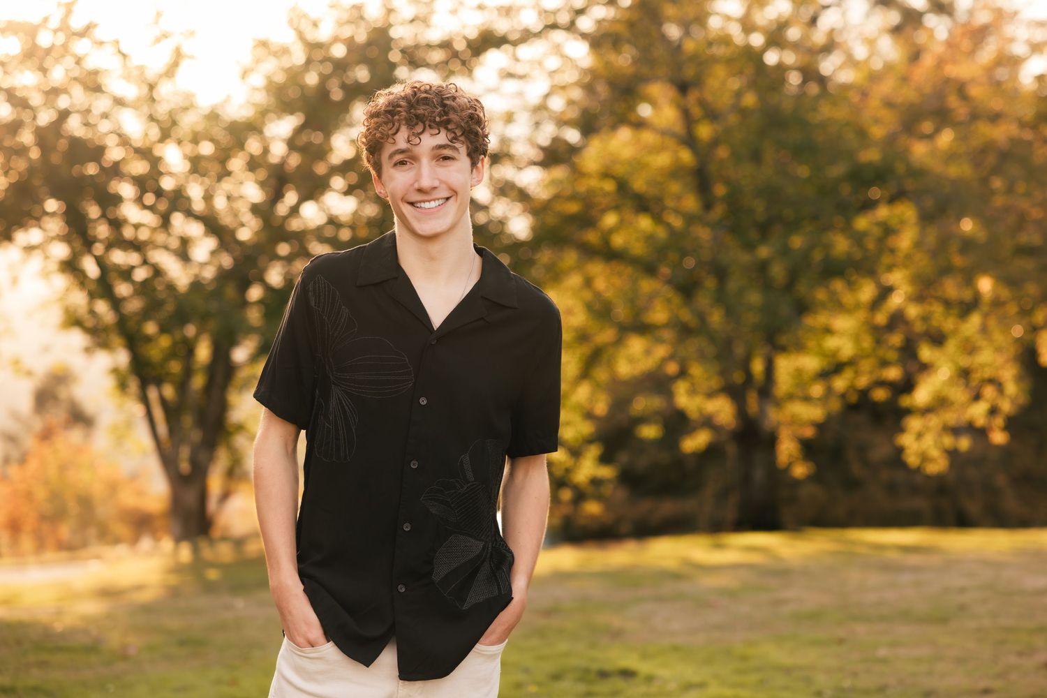 A person in a black button-down shirt stands outdoors in a park setting with autumn trees in golden sunset light.