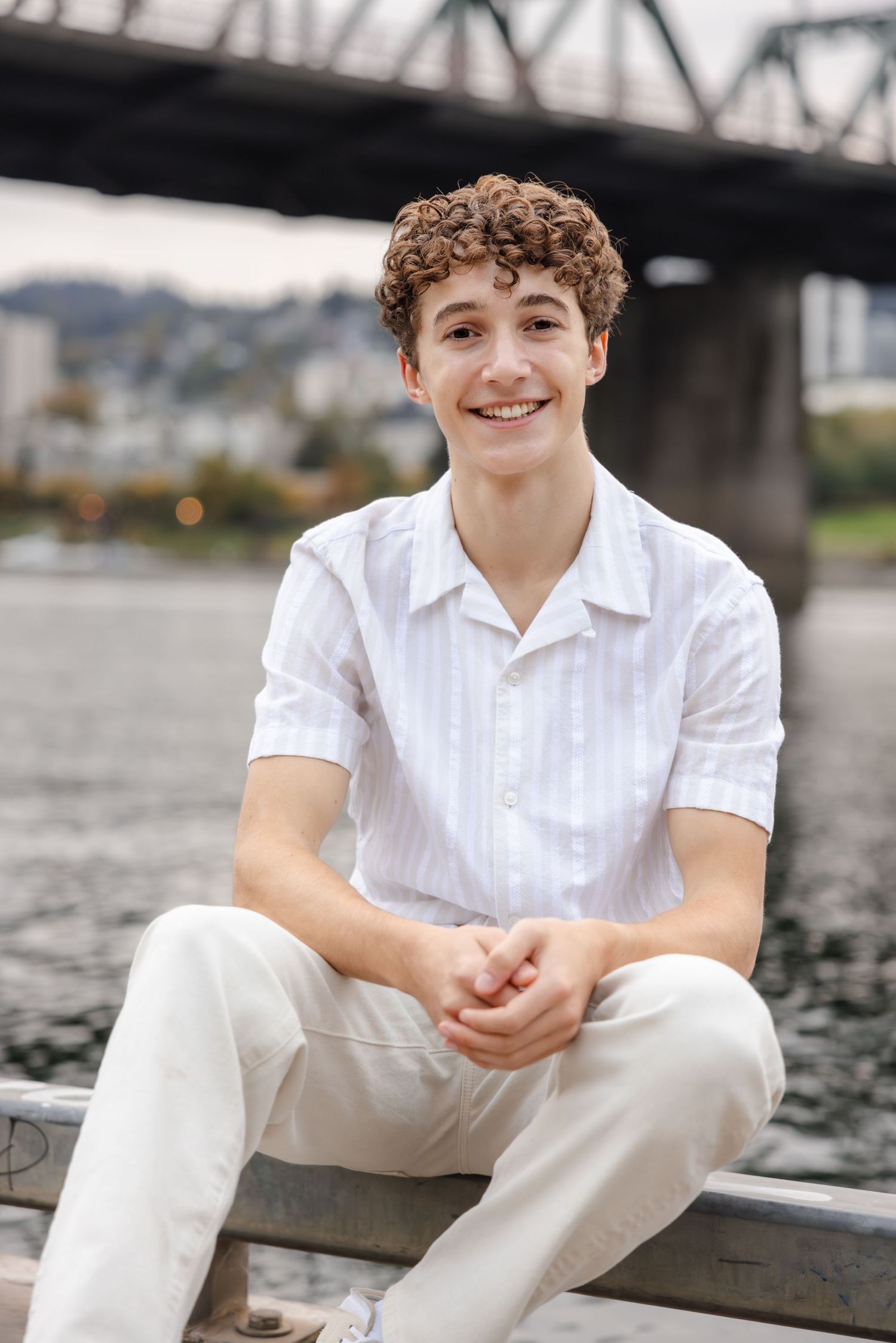 A person wearing a white button-down shirt and light pants sits casually by a river with a bridge in the background.