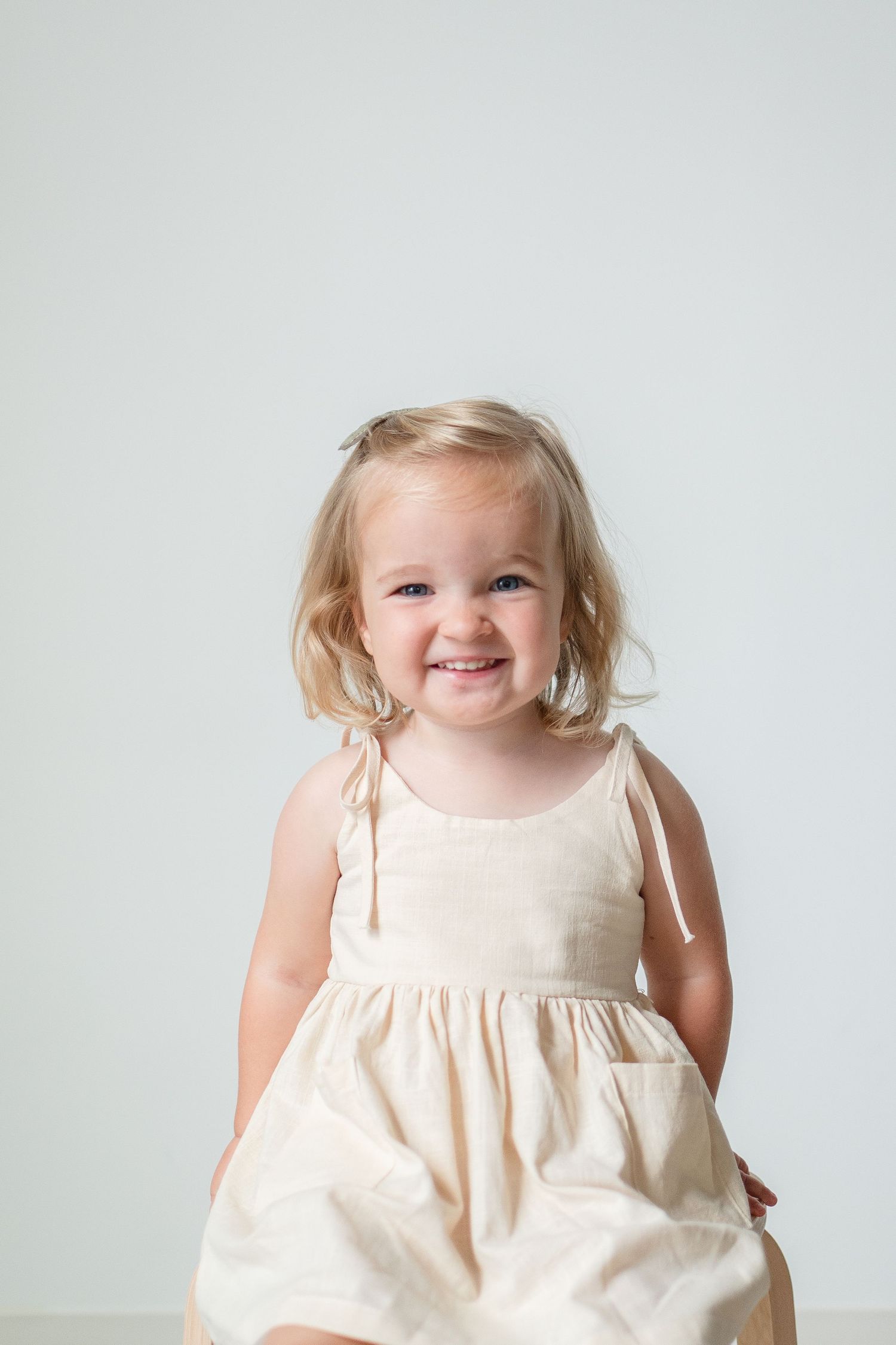 A child wearing a cream-colored sleeveless dress smiles while posing against a plain white background.