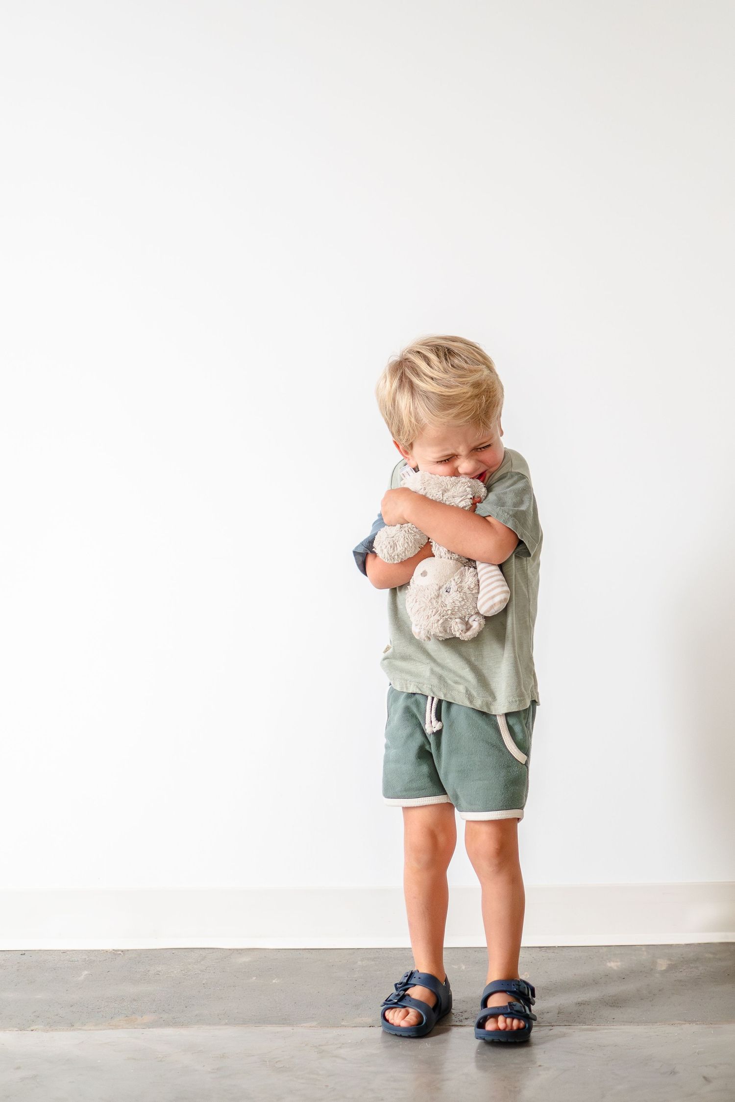 A small child in sage green clothing hugs a plush teddy bear while wearing navy sandals against a white wall.
