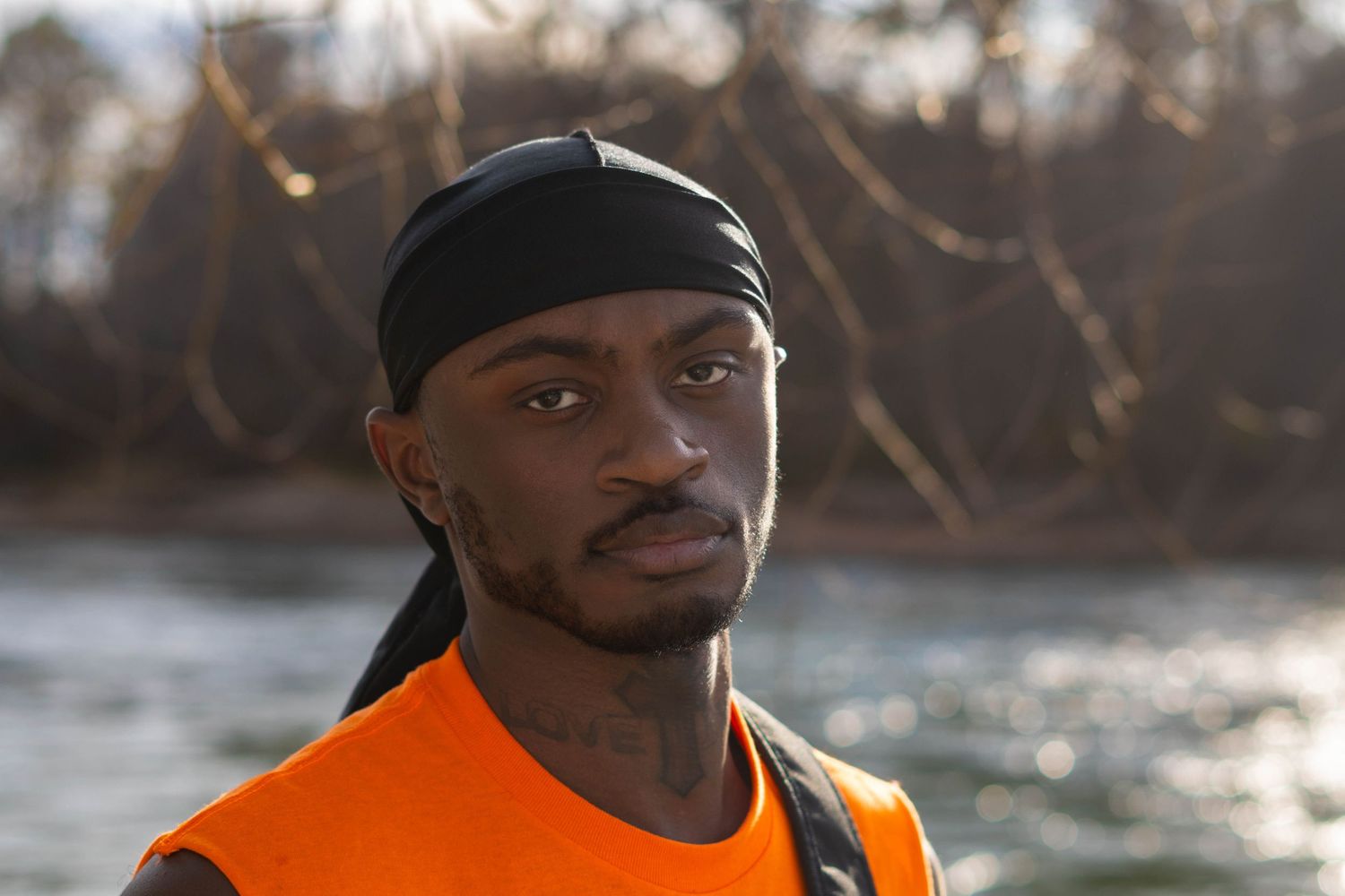 Person in orange safety vest and black durag photographed near riverside in natural lighting.