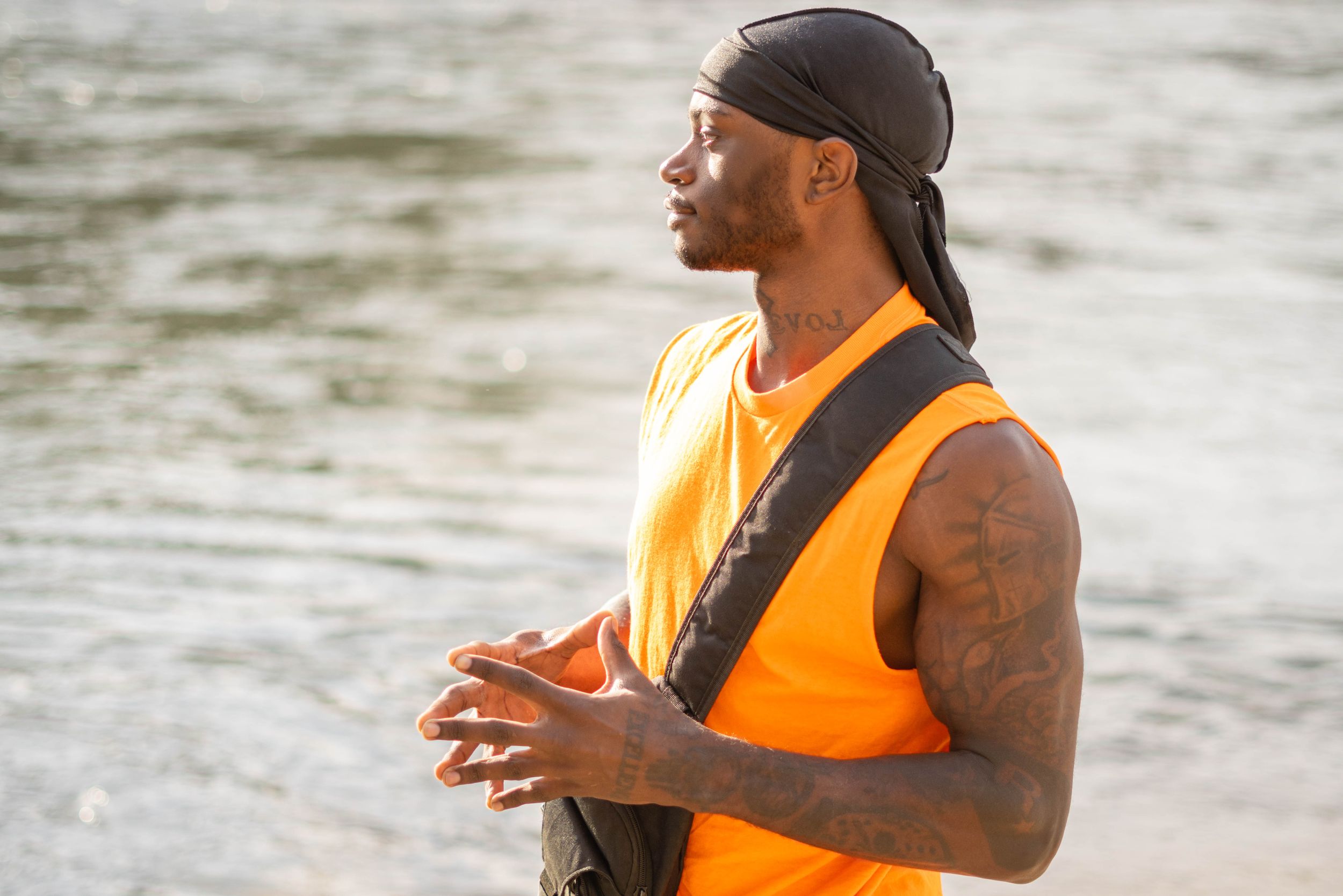 Person in bright orange vest and black durag gazing across water with hands clasped.