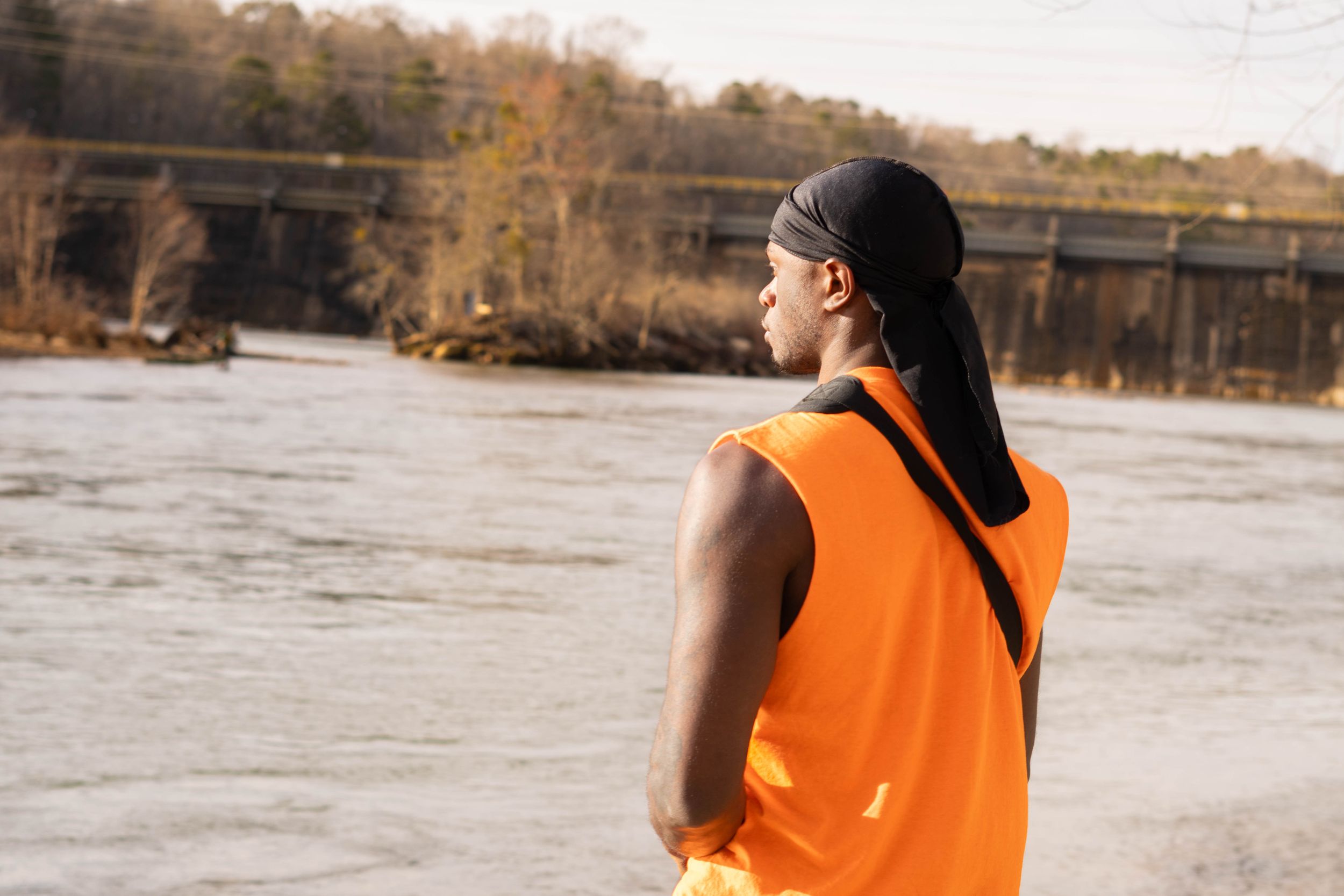 Back view of person in orange vest standing beside river with bridge in background.