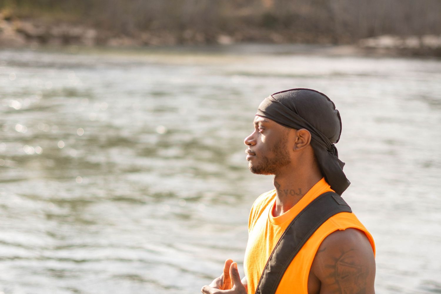 Person in orange safety vest looking thoughtfully across river wearing black headwrap.