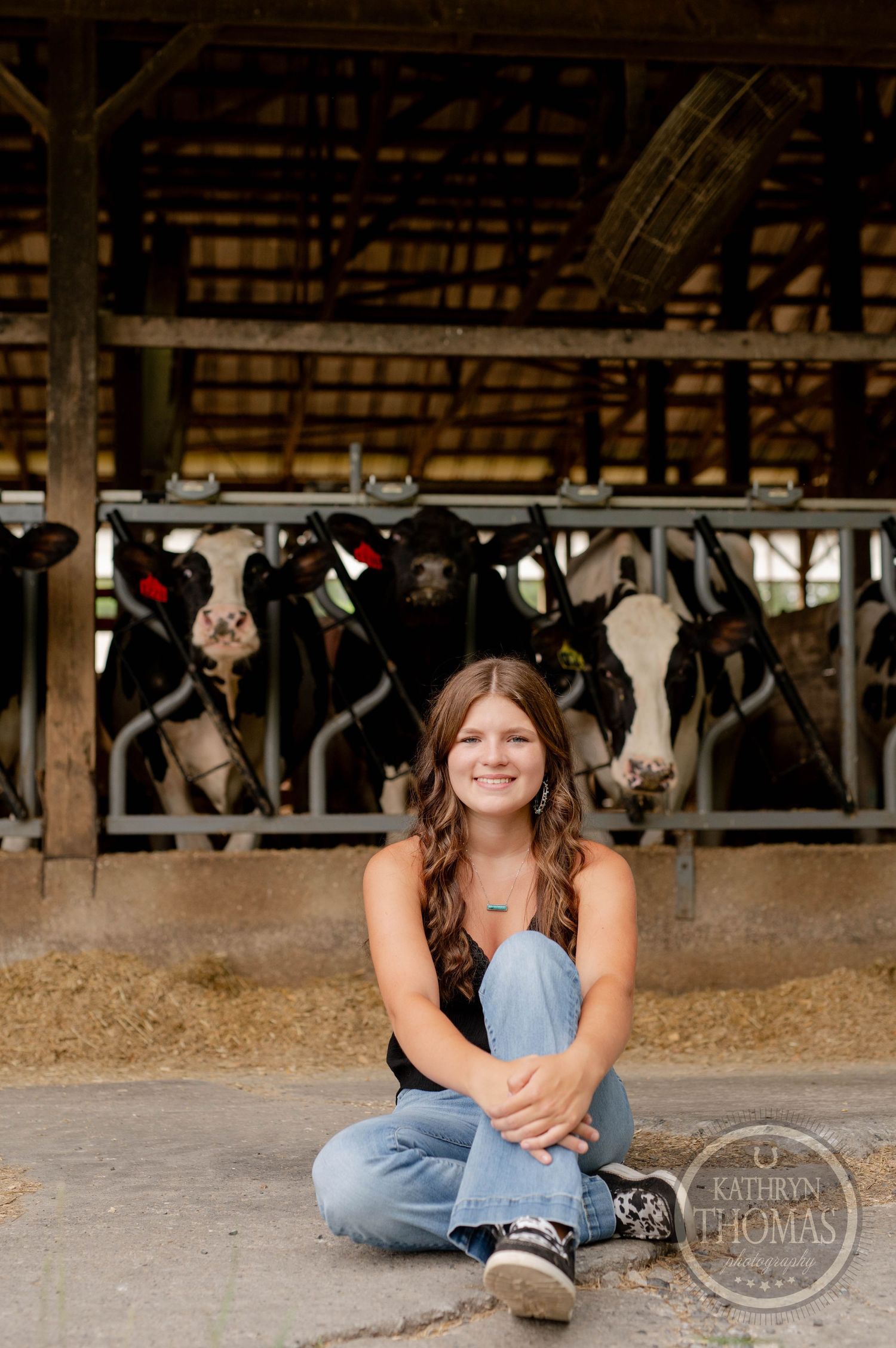 Seated portrait on barn floor with curious dairy cows peeking through stalls in background.