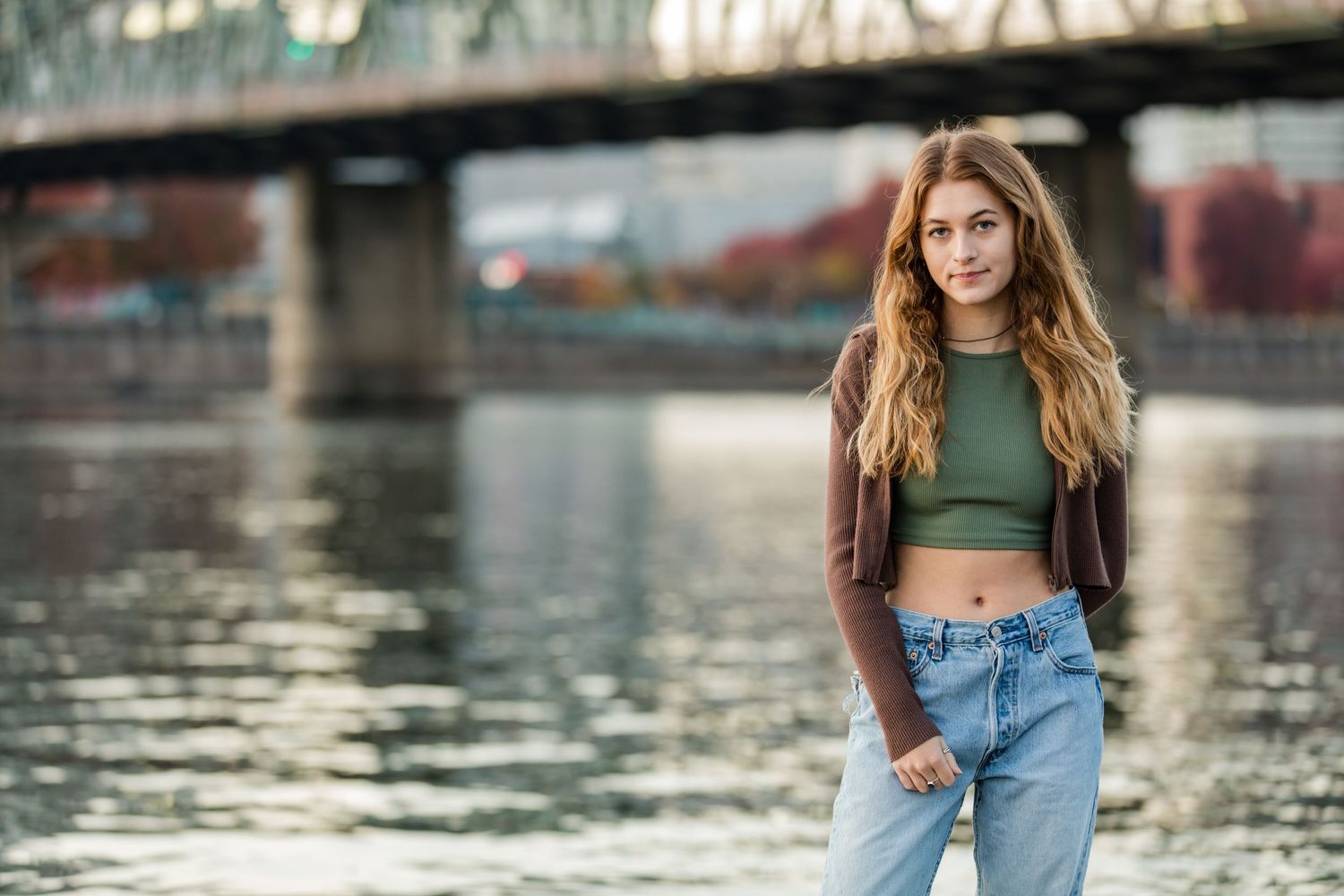 A woman in a green crop top and leather jacket poses near a river with a bridge in the background at sunset.
