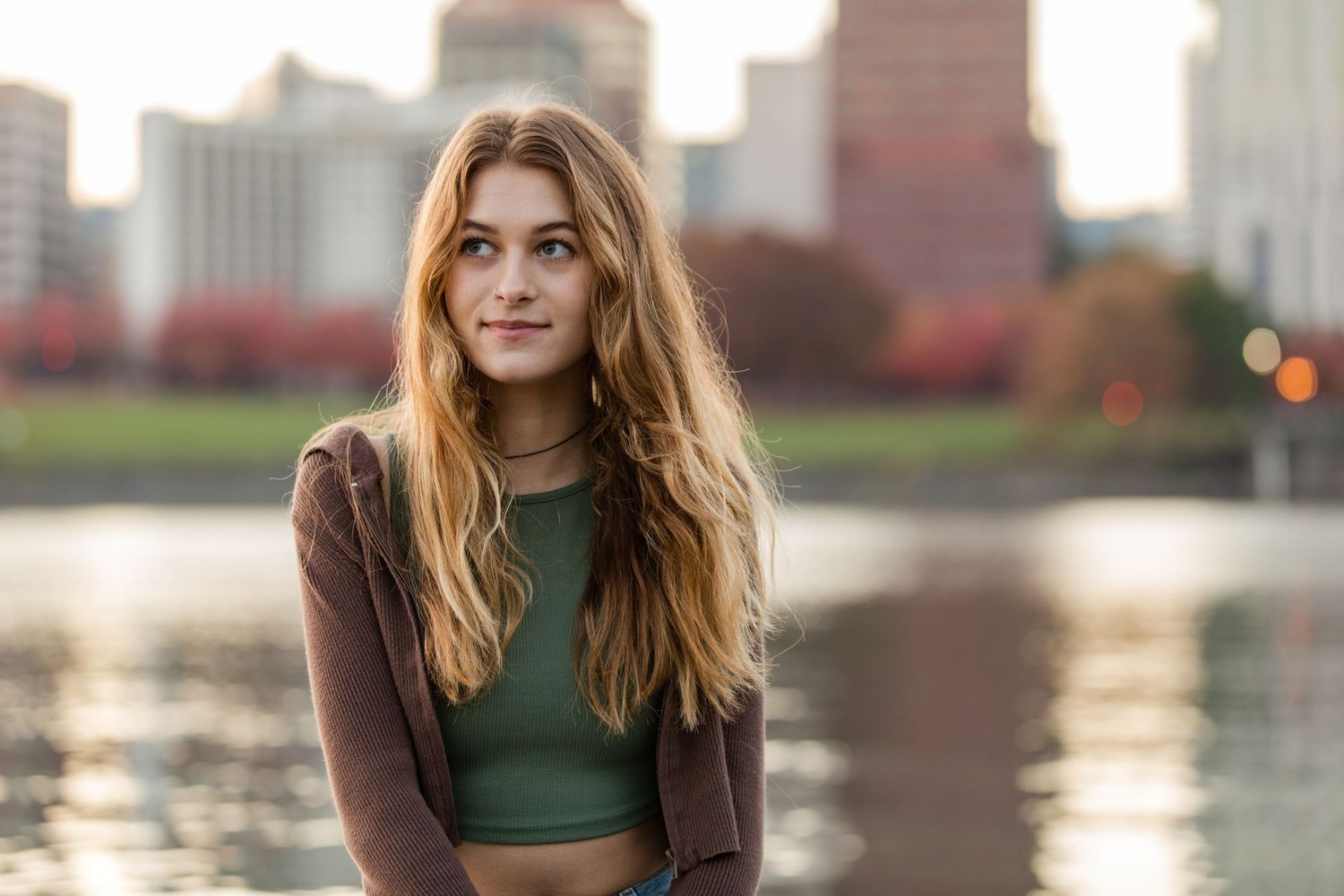 Person wearing a green crop top and brown cardigan poses by a lake with city buildings in the background.