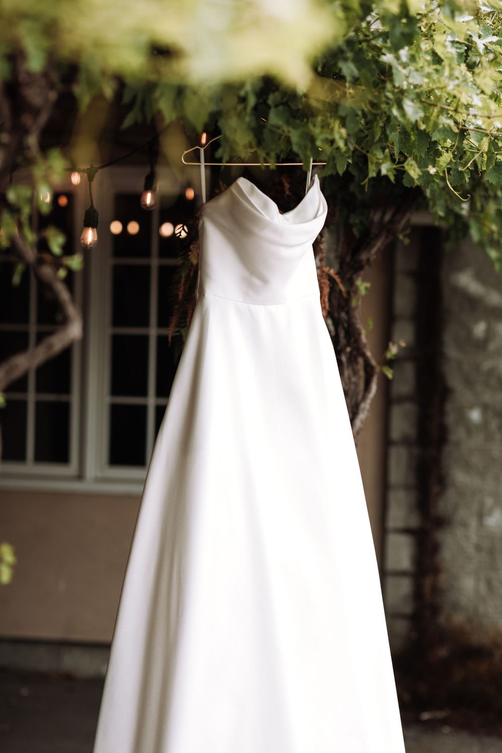 Elegant white wedding dress hanging outdoors with twinkling lights and greenery in the background.