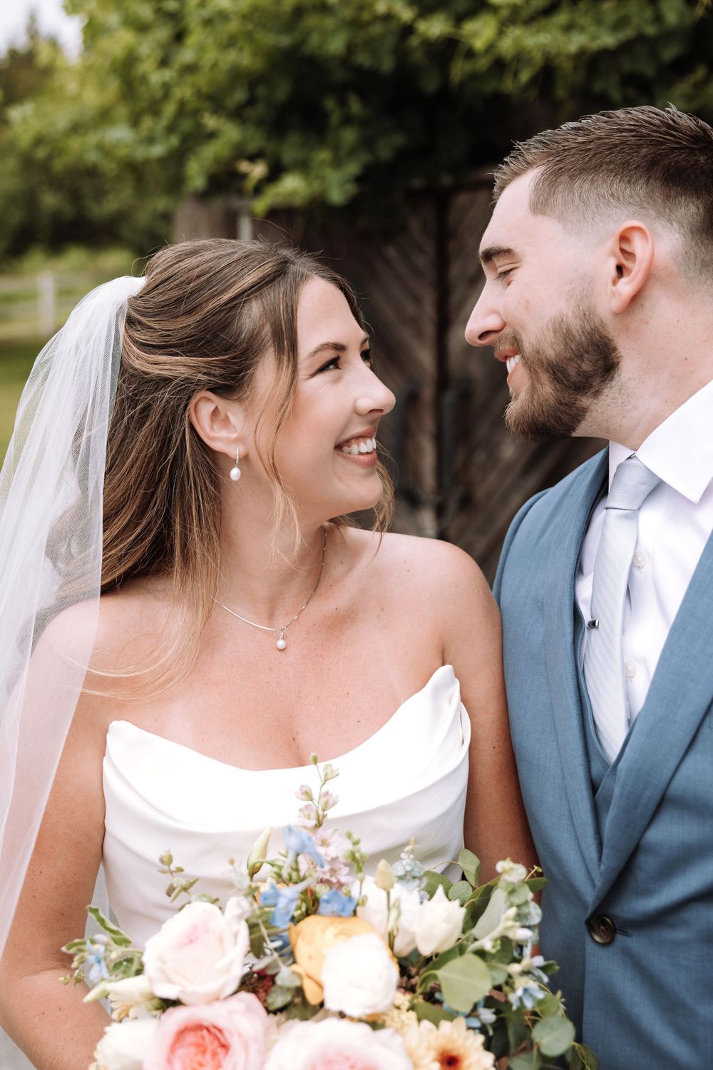 Newlywed couple shares a joyful moment while the bride holds a soft pastel bouquet in an outdoor garden setting.