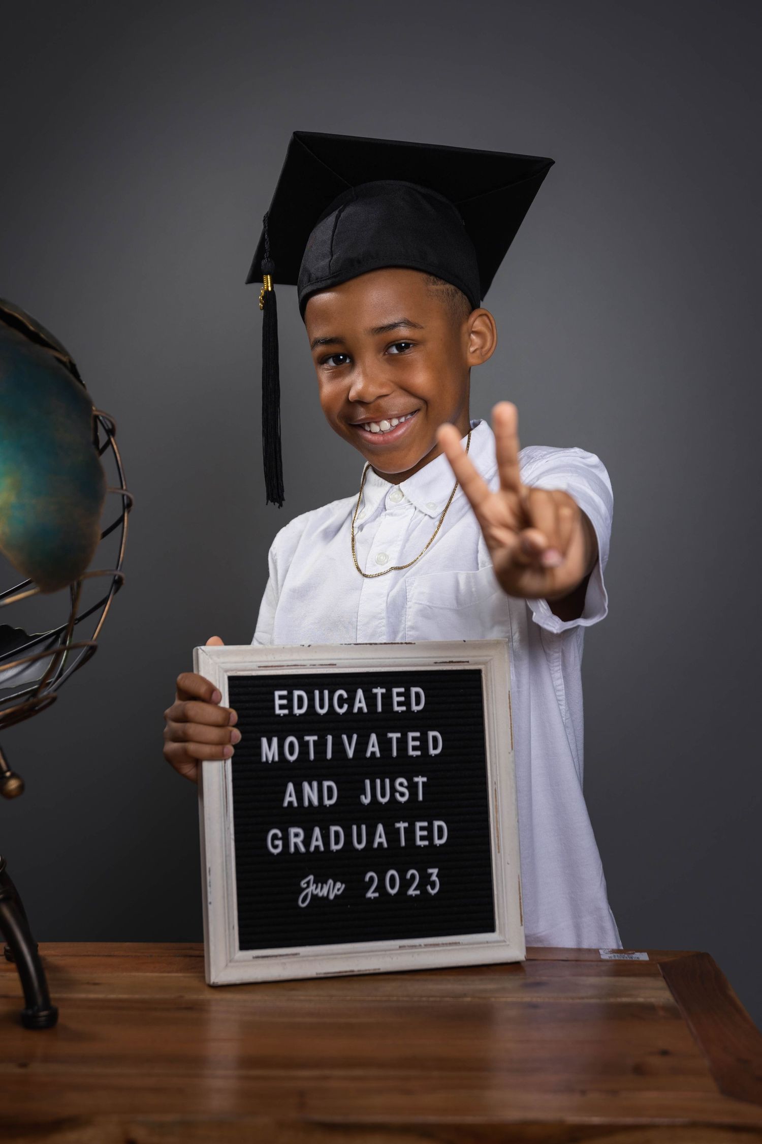 Graduate in cap and white shirt shows peace sign while holding letter board with graduation message near globe.