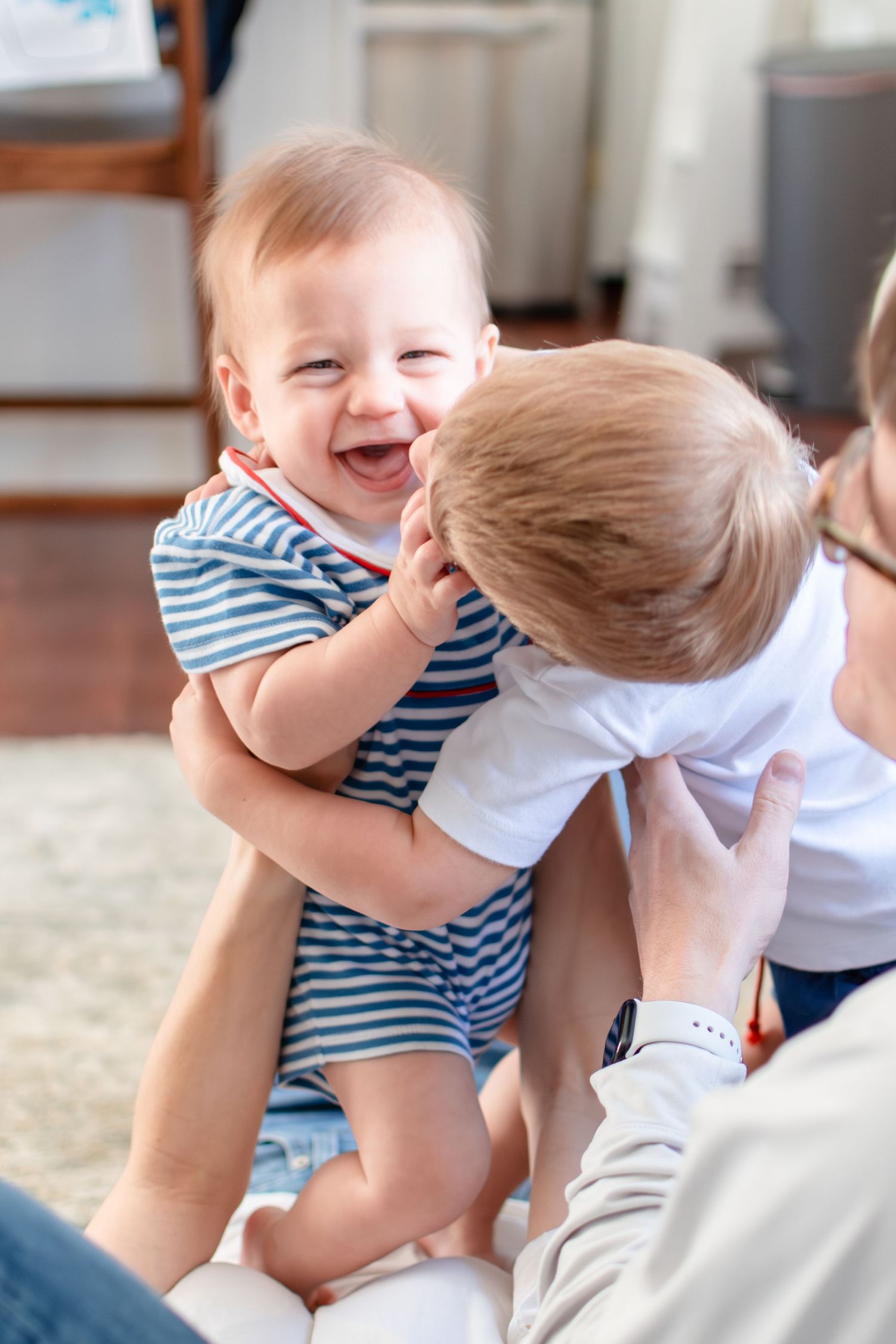 Two small children in playful interaction share a joyful moment while wearing casual clothes on a carpeted floor.