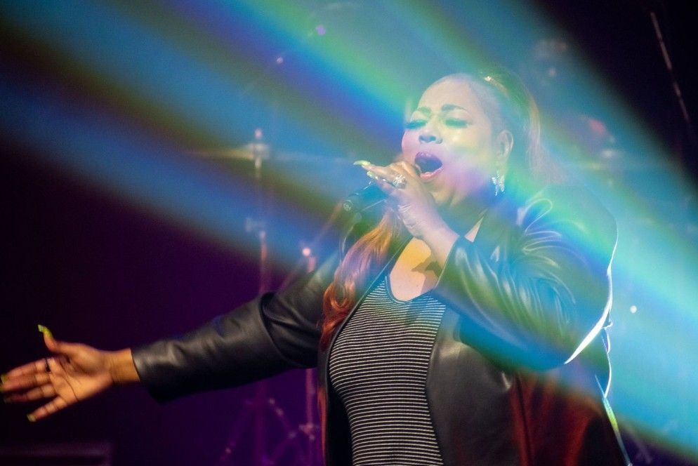 A performer sings passionately on stage with colorful blue lighting and dramatic arm gestures while wearing a leather jacket.