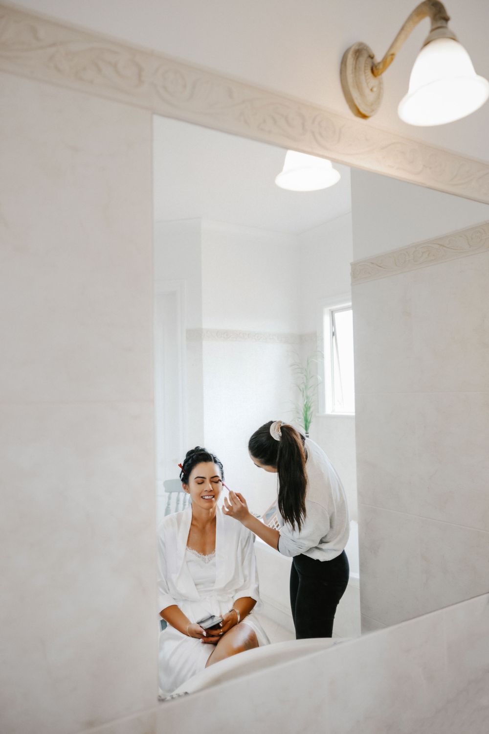 A person wearing a white robe sits in a room while getting their hair and makeup done in preparation for an event.