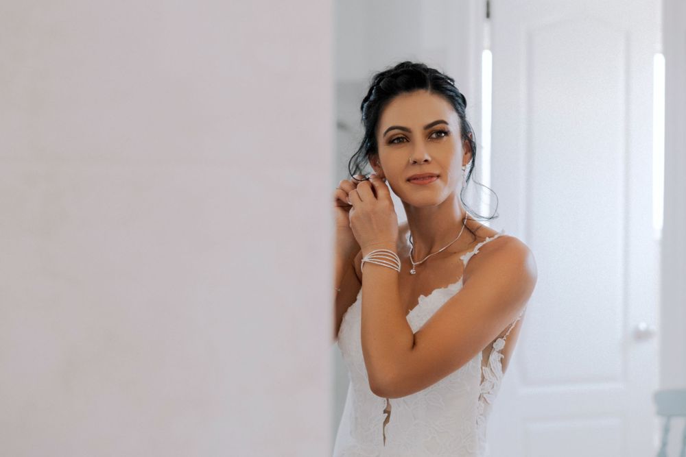 Figure in white dress delicately puts on earrings while getting ready near a window with soft lighting.
