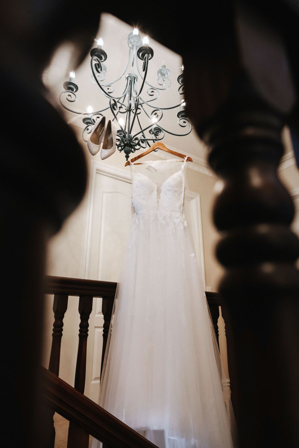 An elegant white wedding dress hangs beneath an ornate chandelier on a staircase in soft, dramatic lighting.