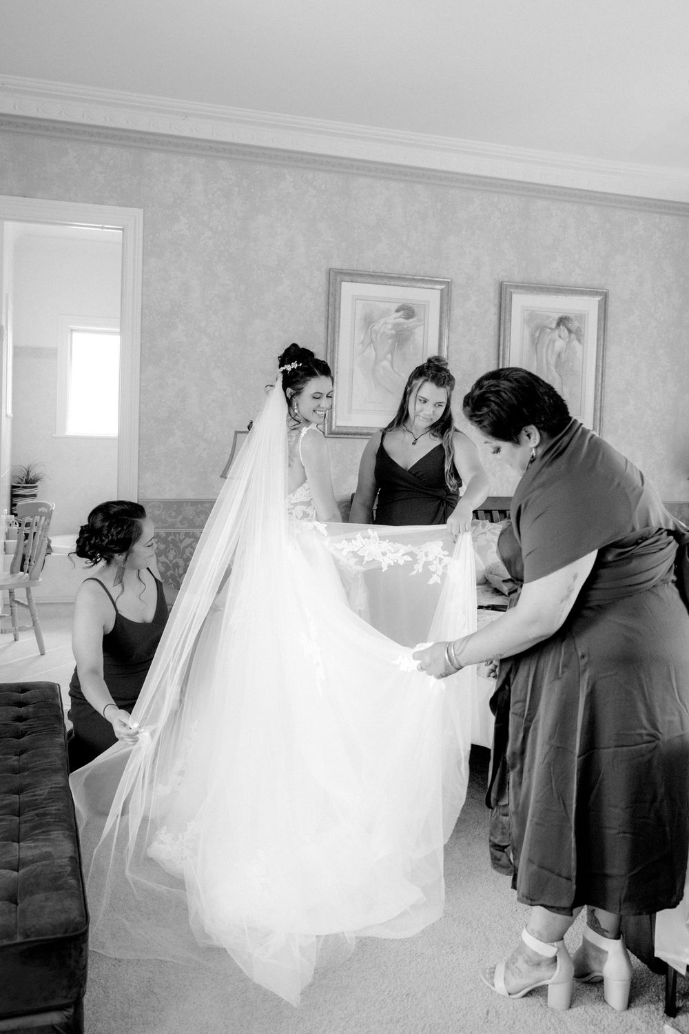 Black and white photograph showing final preparations of a wedding dress and veil in an intimate room setting.