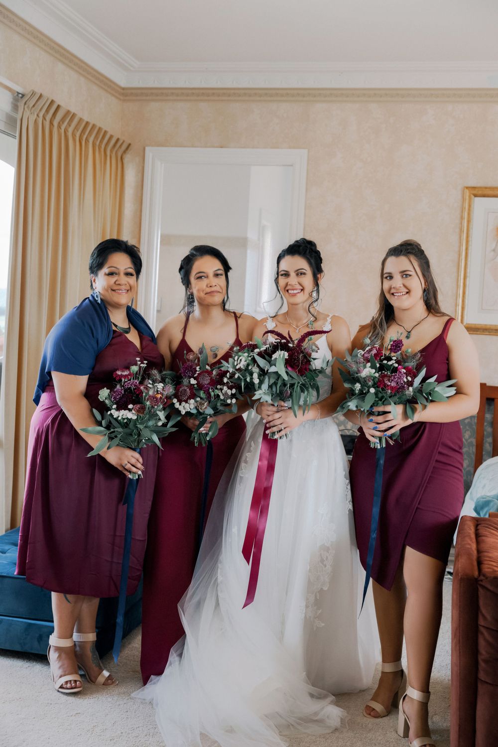A wedding party poses together holding burgundy and pink floral bouquets in a hotel room.