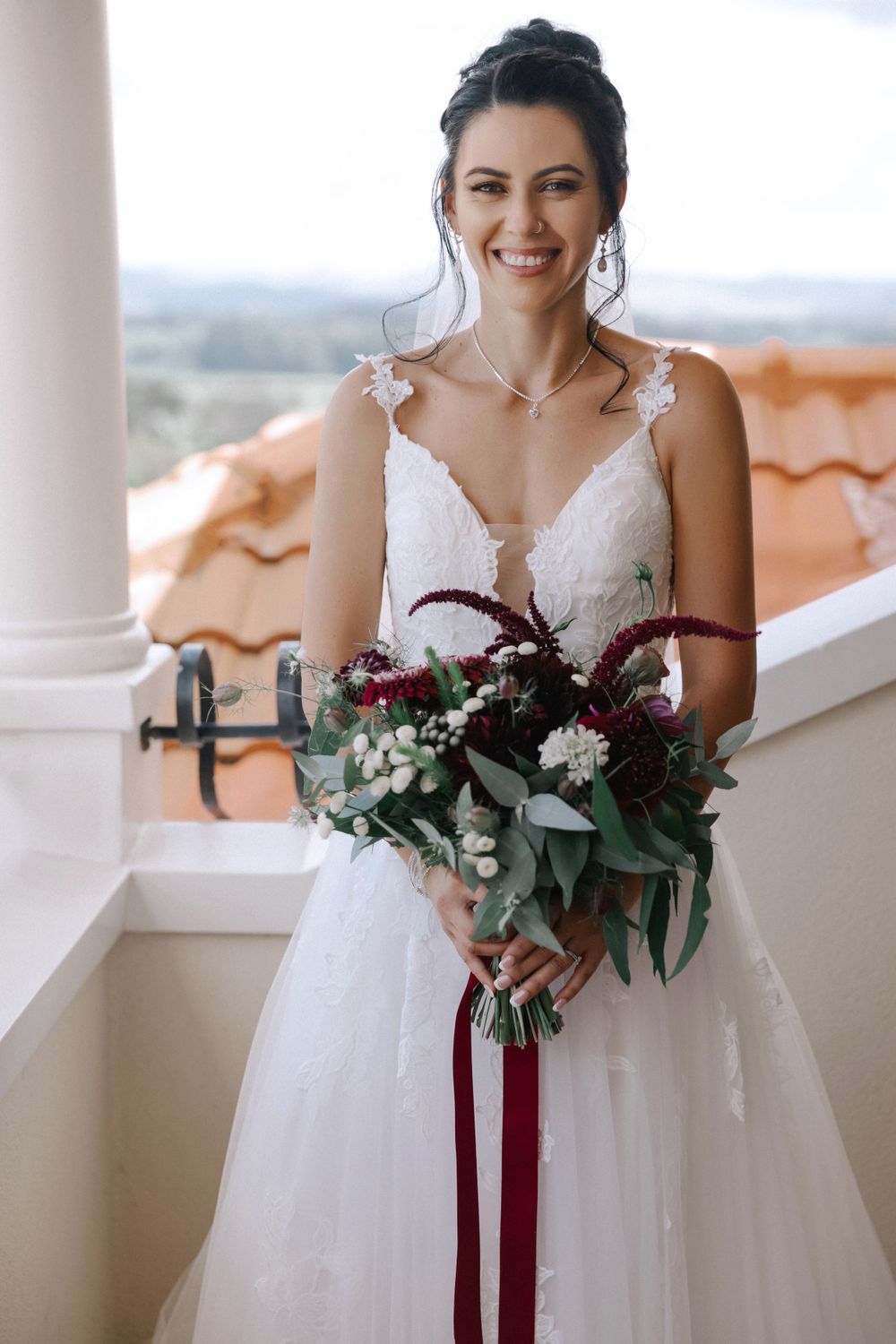 A bride in a white lace gown holds a romantic burgundy and eucalyptus bouquet on a balcony with terracotta roof tiles.