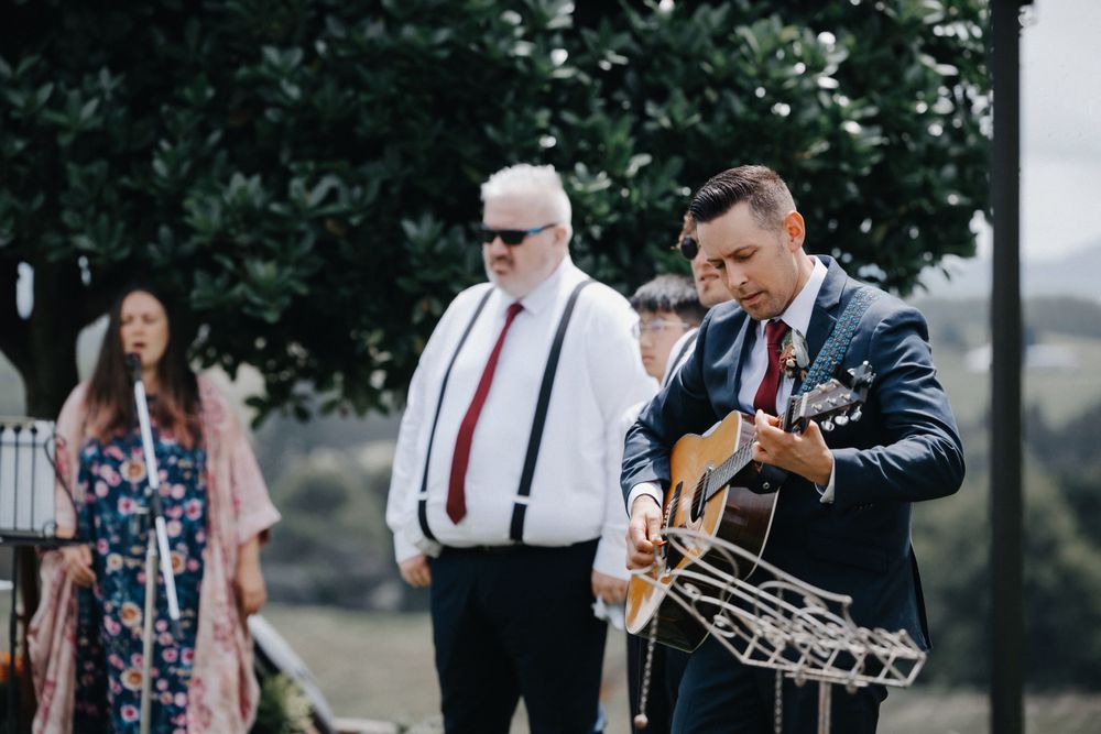 Live acoustic guitarist in navy suit performs at outdoor gathering while guests watch in background.