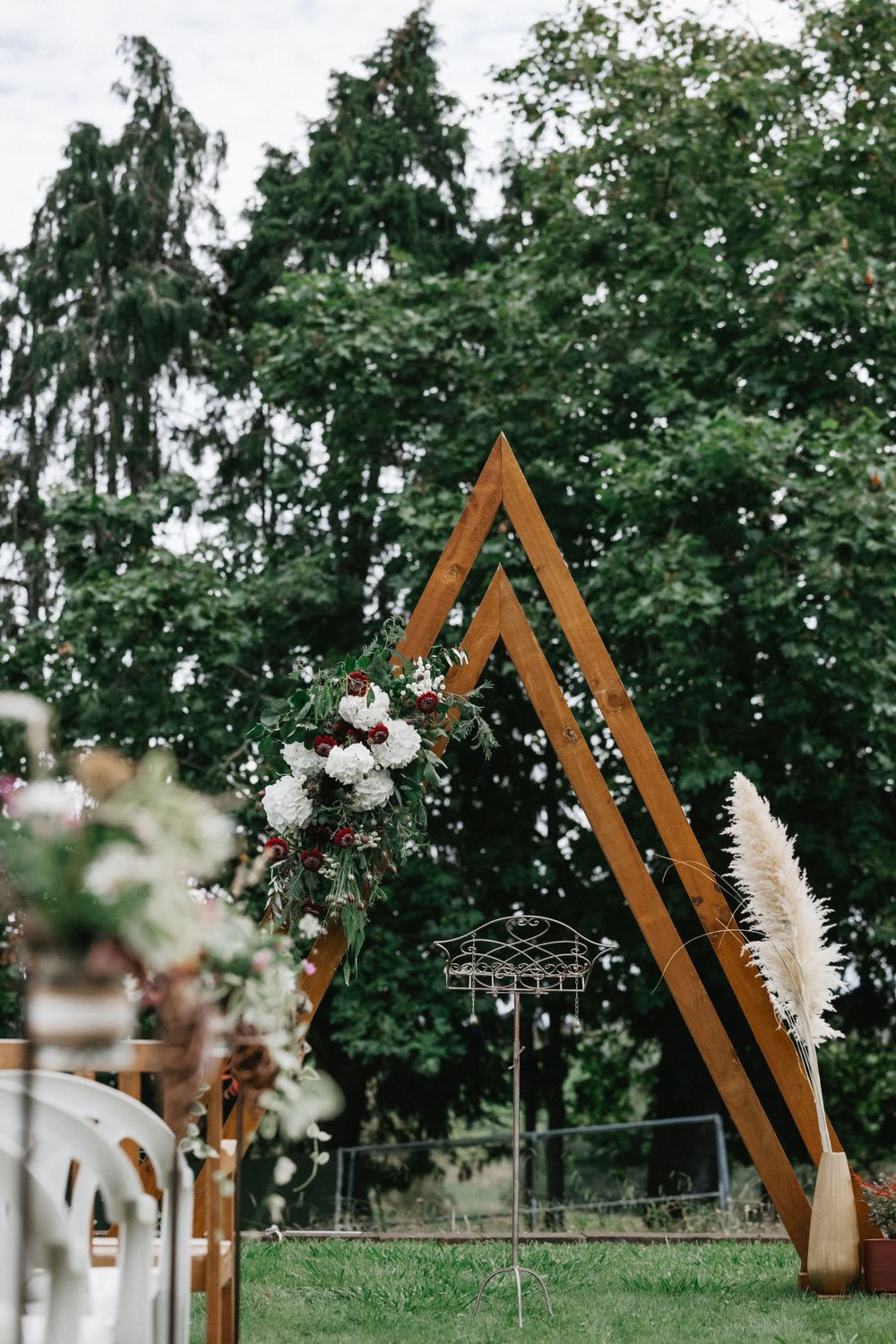 Modern wooden triangle wedding arch decorated with white flowers and pampas grass against green trees in outdoor setting.