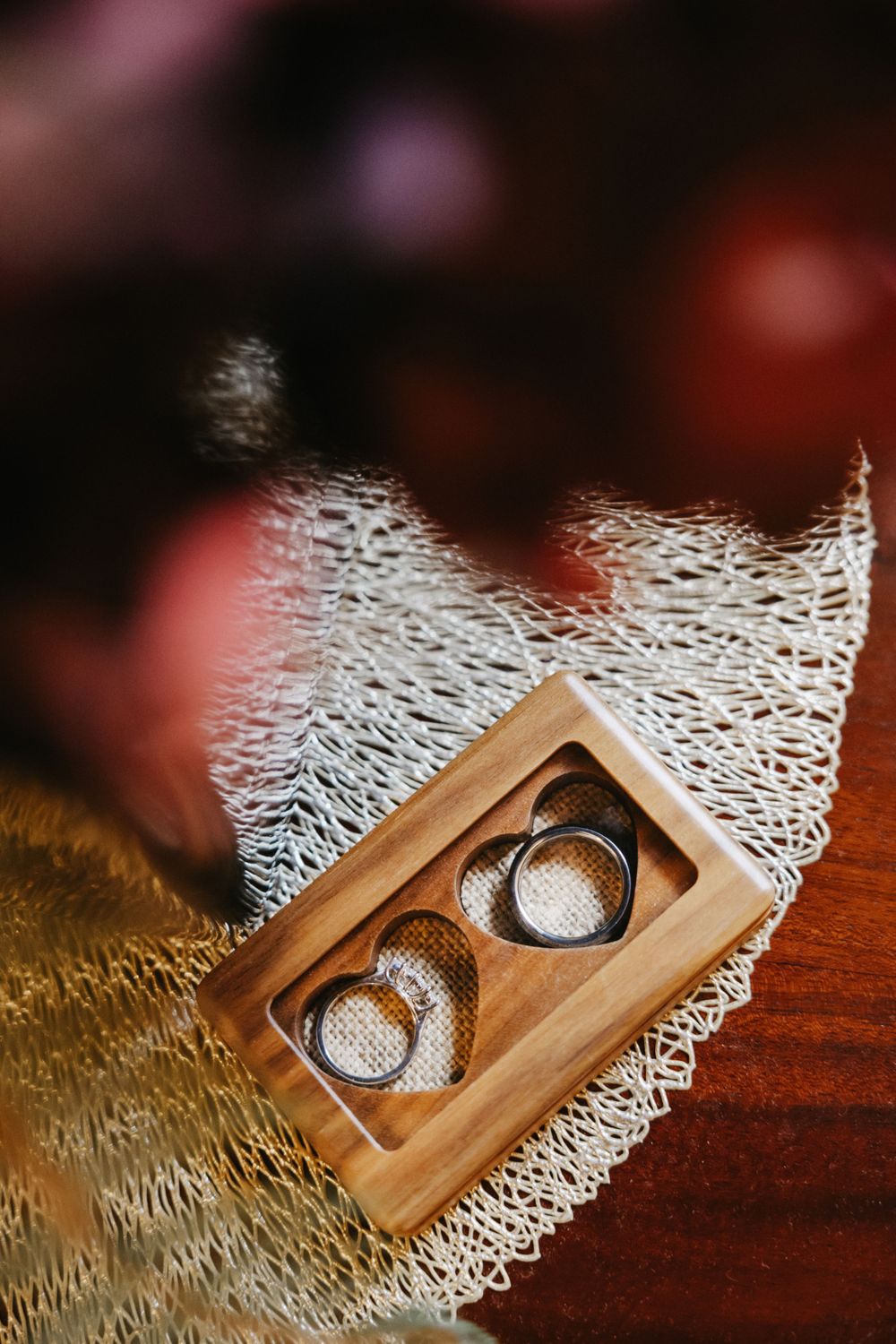 Two wedding rings rest in wooden heart-shaped box on delicate white lace doily against dark wood surface.